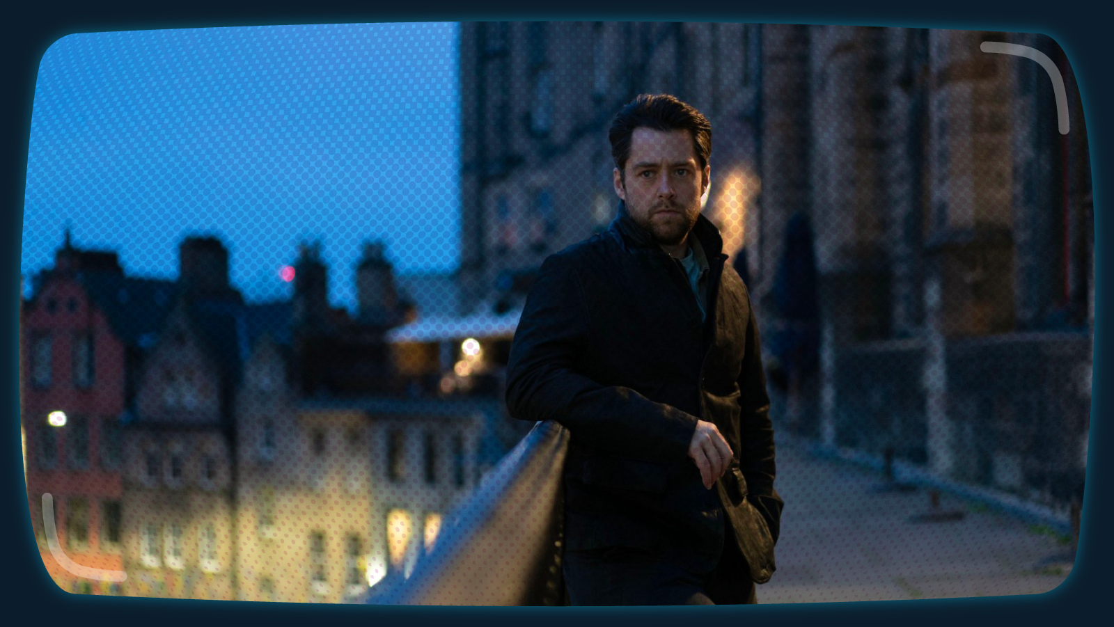 A man in a dark coat leans casually against a stone railing on a city street at dusk. The background is blurred, showing old buildings with warm lights glowing in the windows. The sky is a deep blue, and the scene has a moody, atmospheric feel.