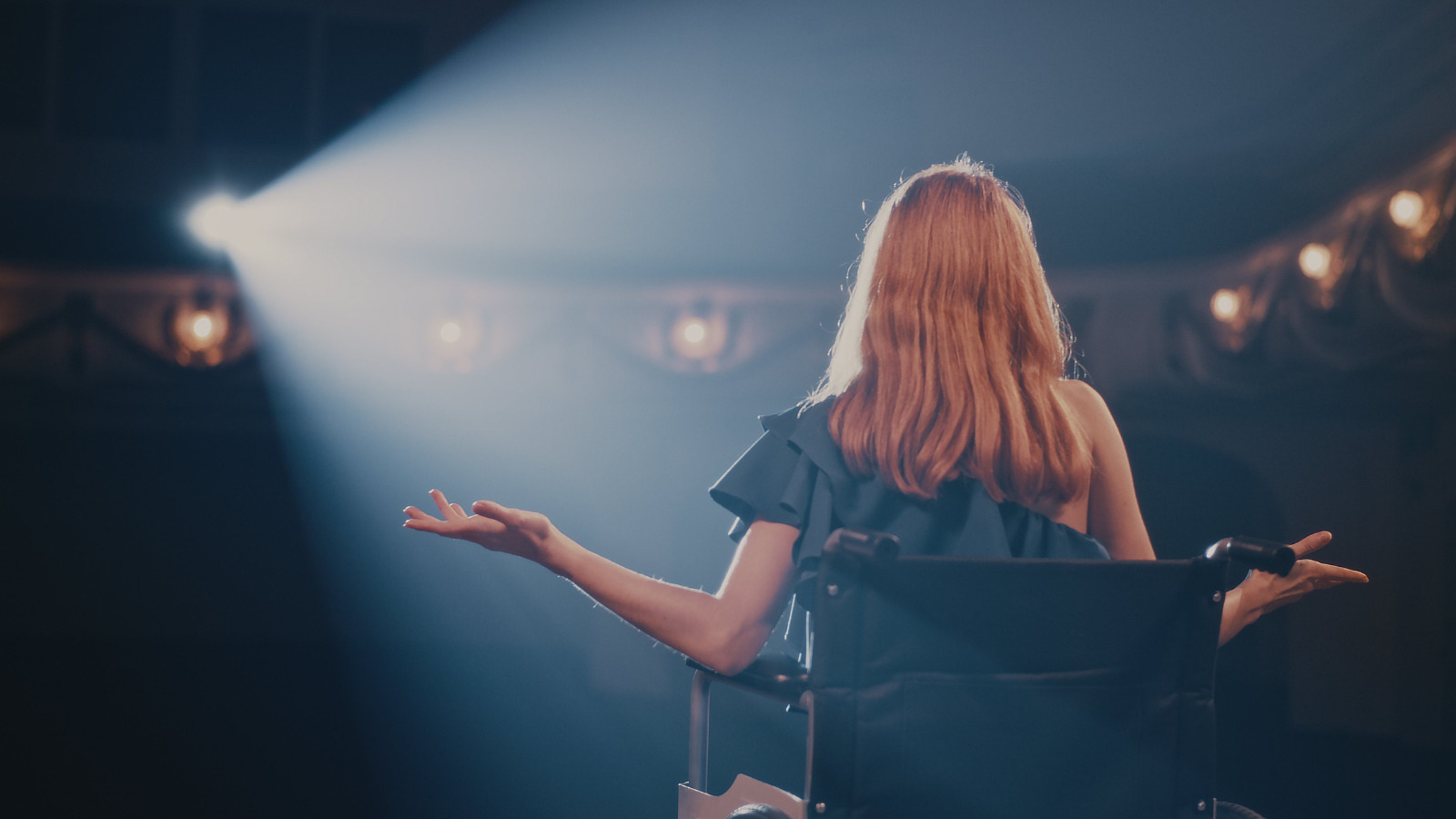 A theatre spotlight lights up the silhouette of a woman with long blonde hair in a fancy blue dress, sitting on a stage in a wheelchair