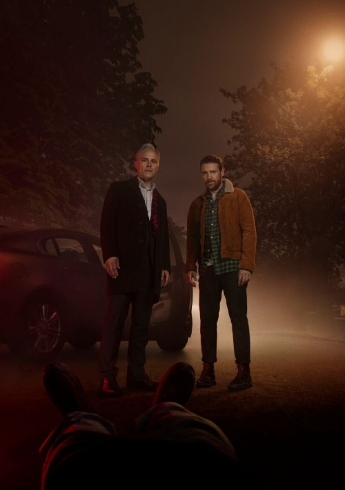 A dramatic image of two men looking at a body lying in the road on a dark night