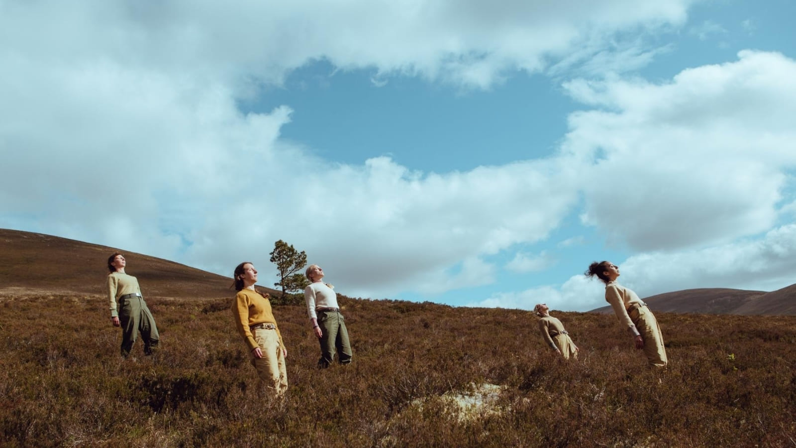 On a hillside against a blue sky, five people spread around in the foreground stand but lean back as though being blown by the wind