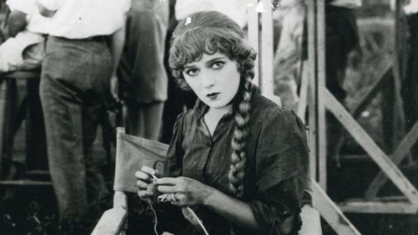 A woman in 1920's attire sits in a chair and looks towards the camera. The image is sepia