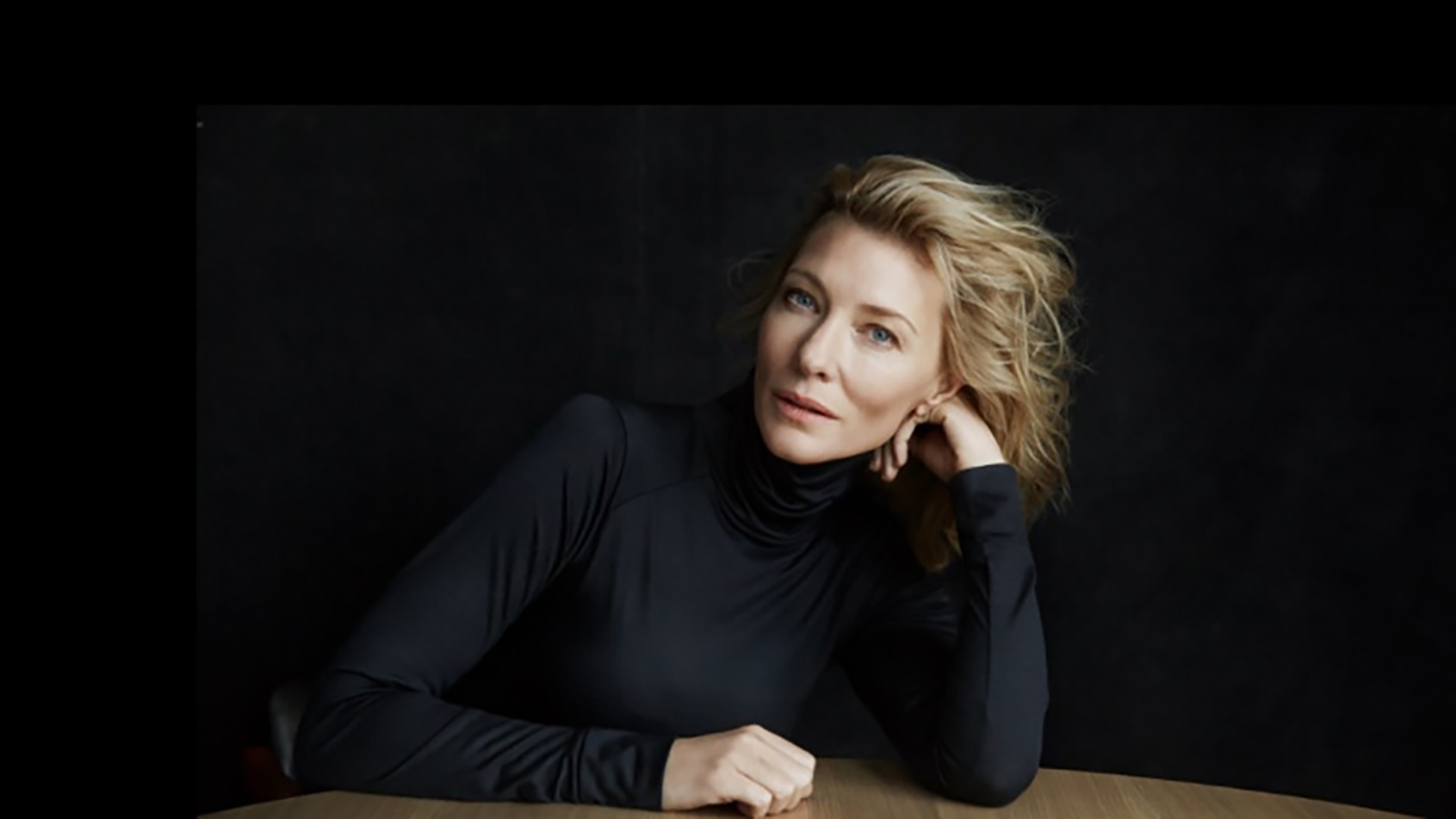 Cate Blanchett with her elbow on a table, wearing a black jumper against a black background