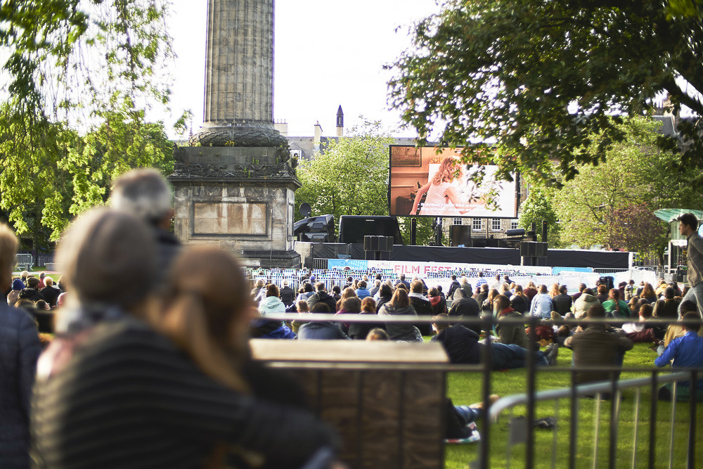 People sitting in st Andrews square in Edinburgh watching a large outdoor screen