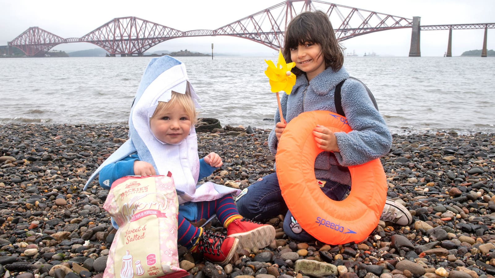 Two children sit on the banks of the Forth with the bridges in the distance behind them