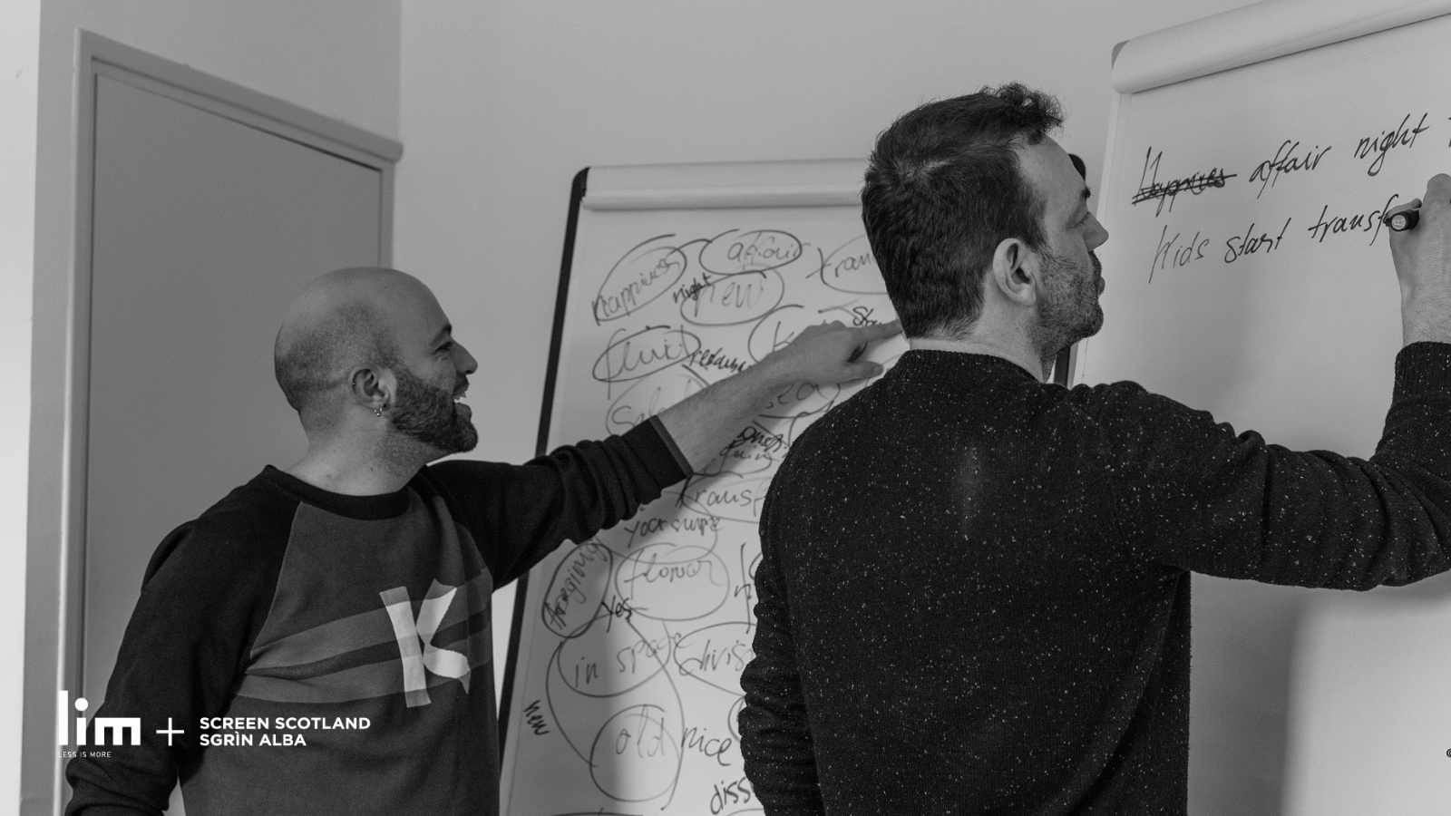 A bald man with a beard in a long sleeved top with the sleeves rolled up points at an obscured word on Flipchart covered in black writing. It looks like a brainstorming session. Another man to the right is writing 'kids start....' on another Flipchart.