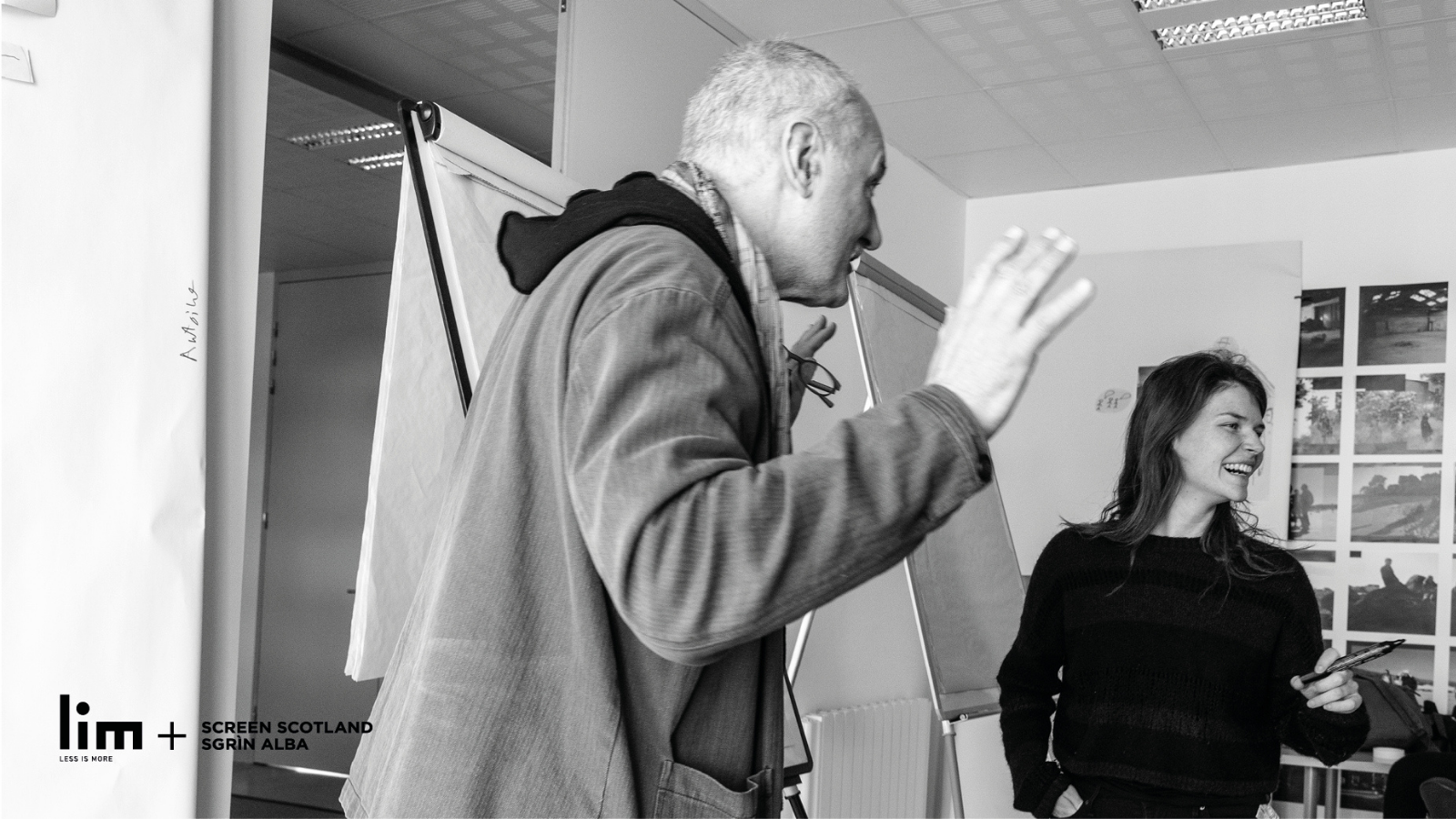 An older man with short hair wearing a hood and jacket raises his hands in a gesture as he talk to someone we can't see. A woman next to him holds a pen and is laughing. Behind them are two blank flip charts.