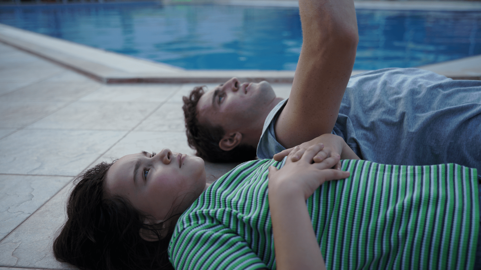 A still from Aftersun - a young girl in a green striped t-shirt lies on the tiled floor surrounding a pool next to an older man in a blue shirt.