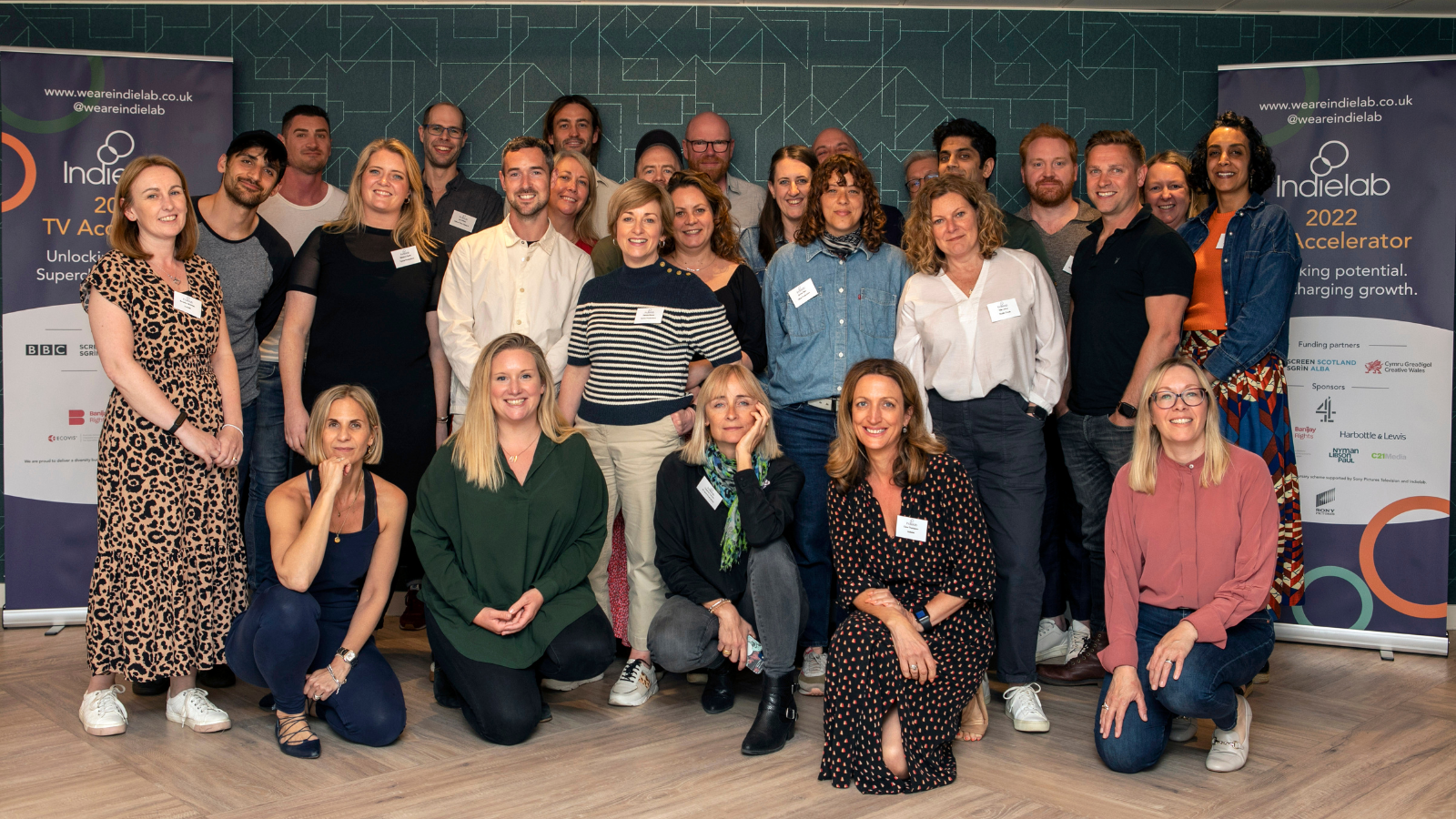 A group of 50 people, some standing, some kneeling, smiling at the camera. On their left and right are banners that read Indielab 2022 Accelerator.