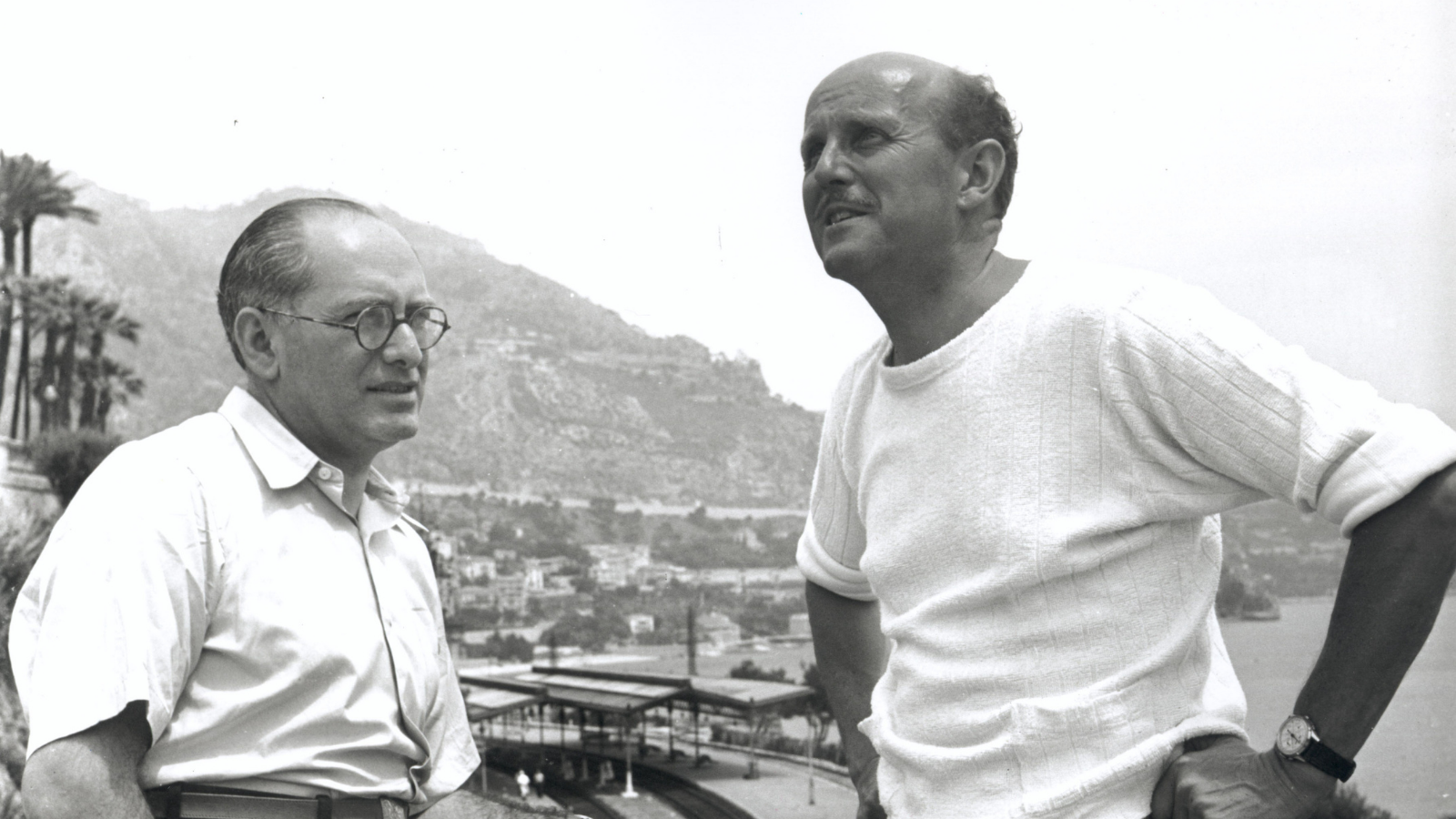 Two men in white shirts, in the distance, the outlines of buildings built into the side of a mountain. There is a harbour to the right. One man looks at the other with a quizzical look on his face, the other is staring off into the distance.