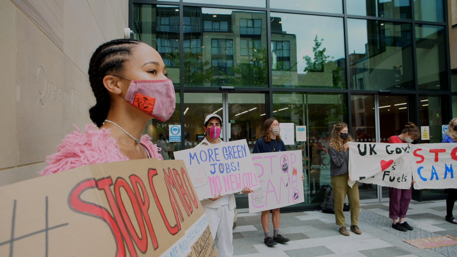 A protest against oil outside a stone built office building. Protestors wearing masks and carrying colourful signs that read 'more green jobs, no new oil', 'stop CAMb' and 'UK Gov loves Fossil Fuel'. This is a still of campaigners from the film The Oil Machine.