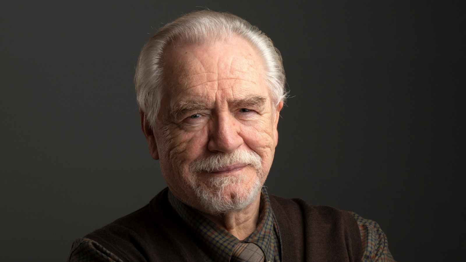An older man with white hair and a beard, wearing a dark shirt. It is Brian Cox. The text reads 