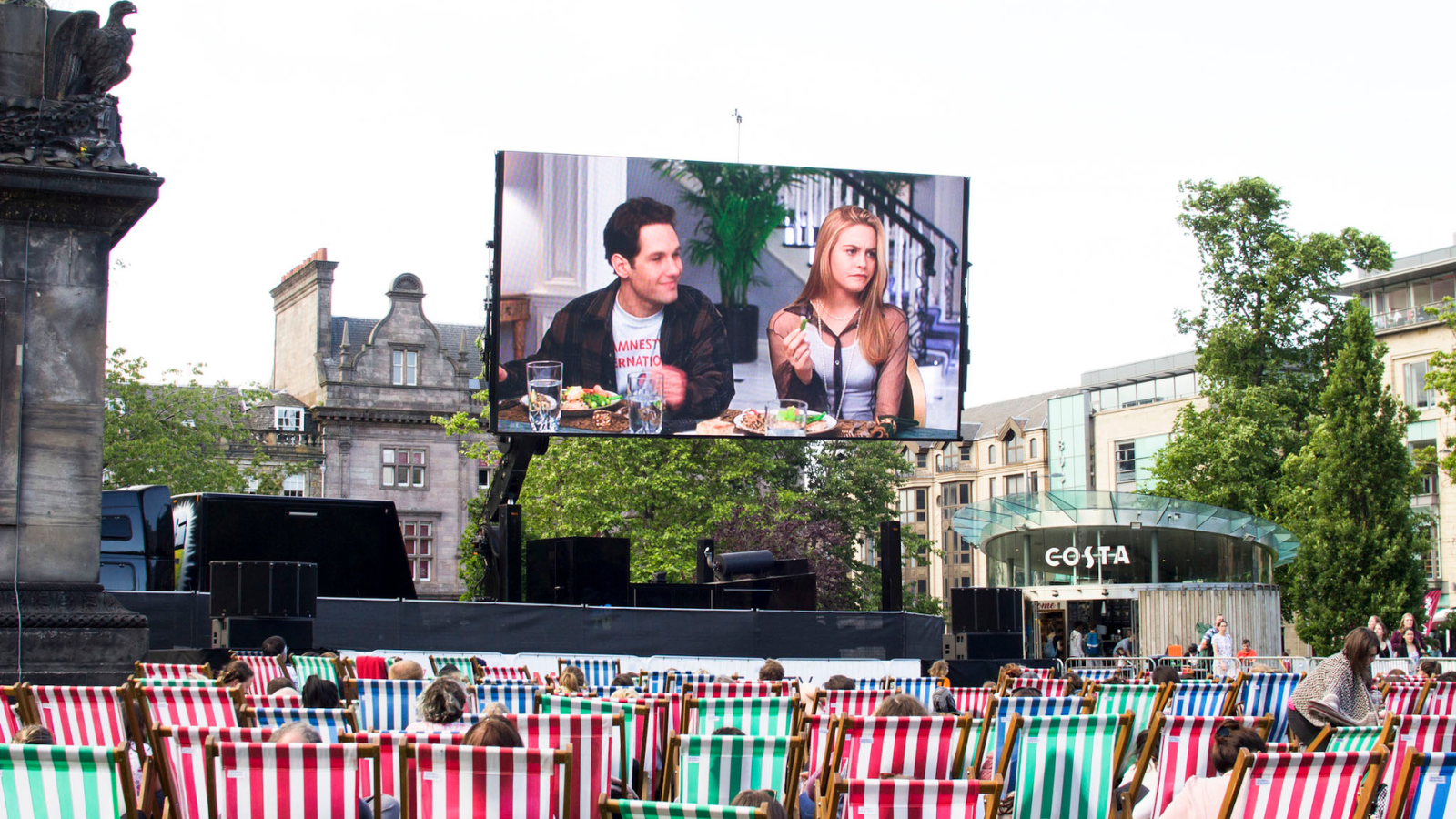 The film Clueless on a large outdoor screen in a leafy park (St Andrew Square) watched by a large crowd of people sitting on the striped beach style chairs during a summer's day.