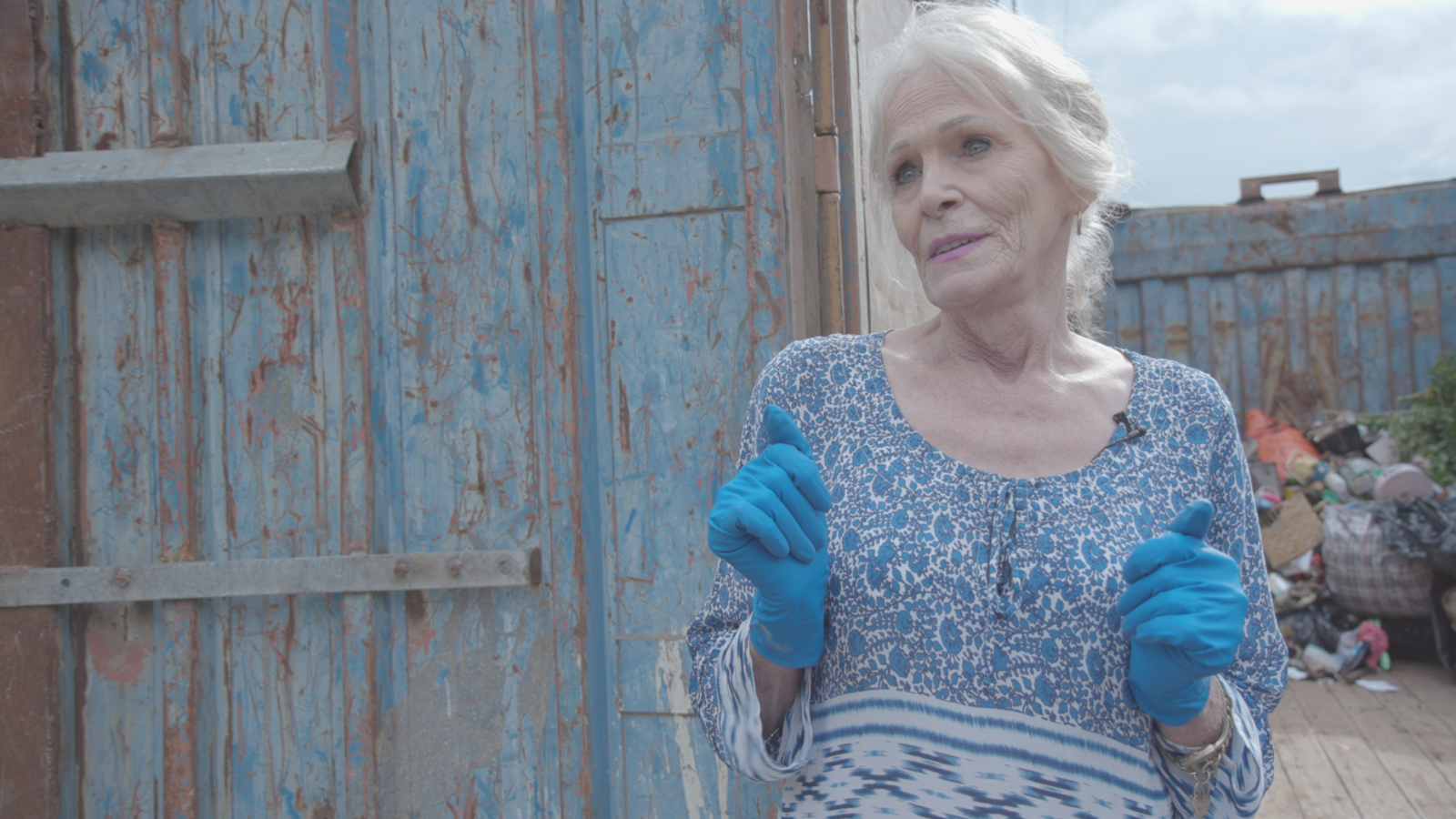 Older woman with white hair and a flowery blue top wearing blue marigold gloves.