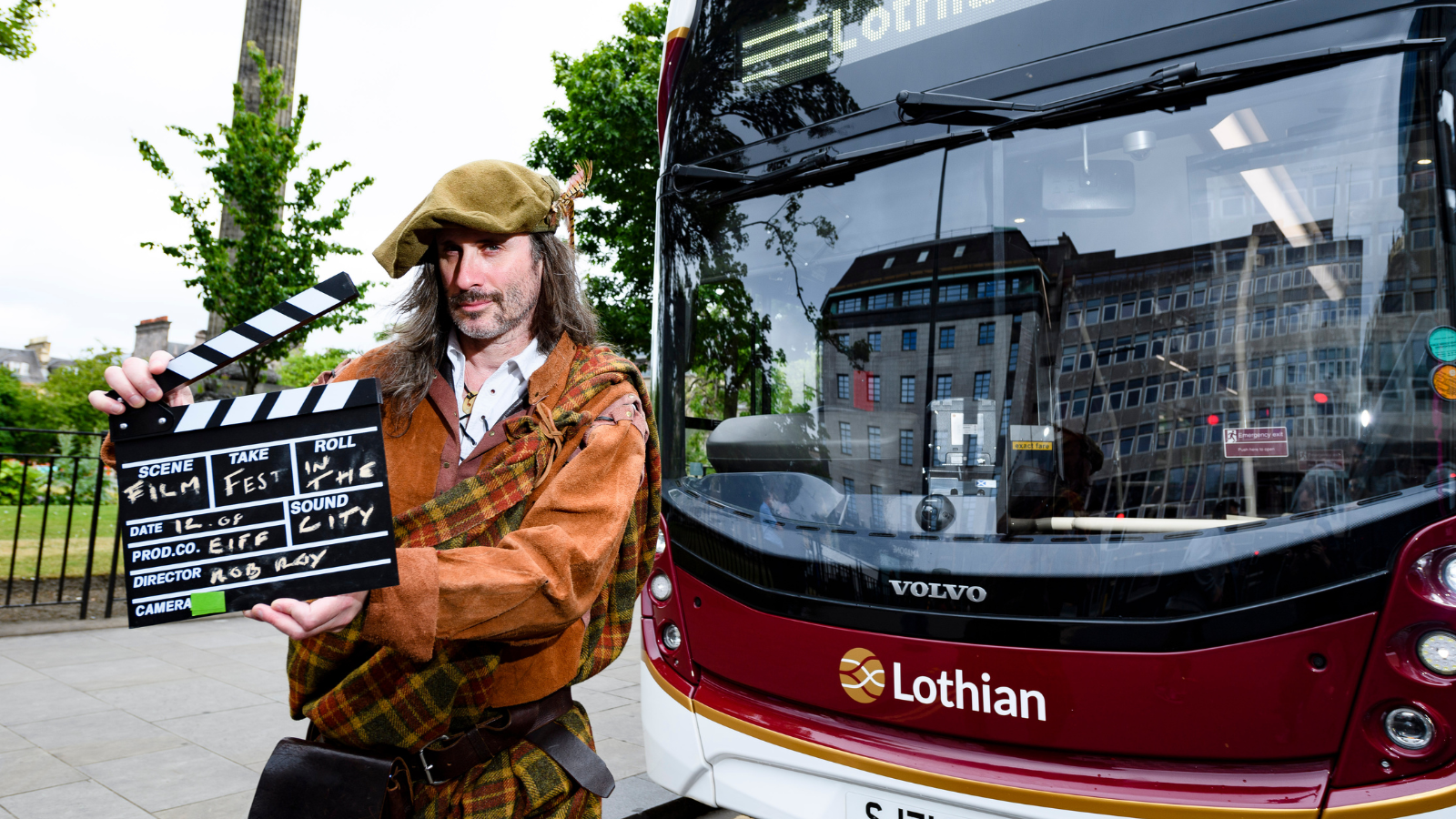 A man in traditional Highland dress holding a clapperboard in front of a red, white and gold Lothian city bus.