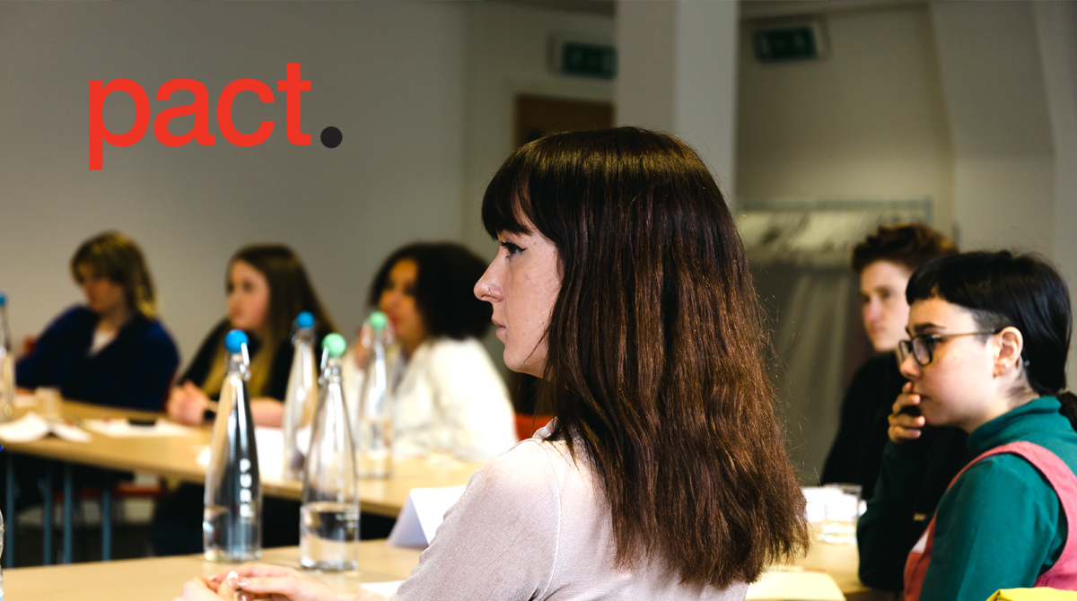 Detail shot of a woman with short black hair and a fringe sitting at a table with other people watching someone out of shot. There are water bottles and notepads on the table. The text reads 'Pact'