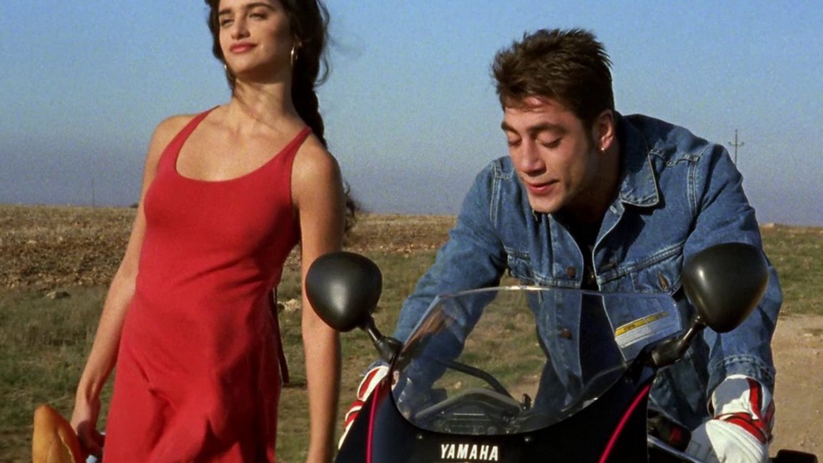 A woman with long dark hair in a red summer dress (Penelope Cruz) and a man with short brown hair in a leather jacket on a motorbike (Antonio Banderas). A still from Jamón Jamón.