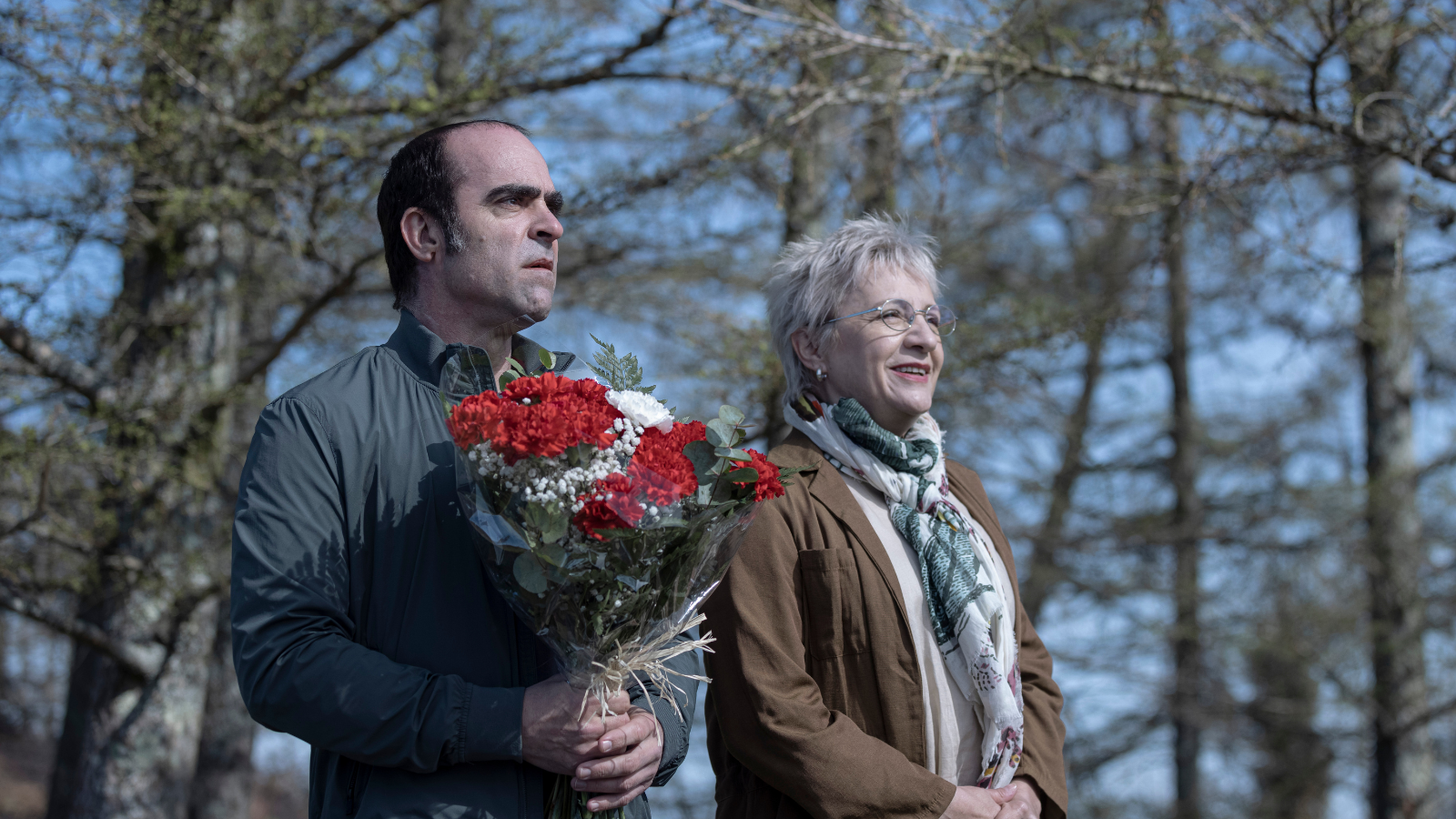 A man with short black hair holding a bouquet of red roses, next to an older woman with short grey hair wearing glasses and a scarf. They are outside on a winter's day, looking out to something in the distance.