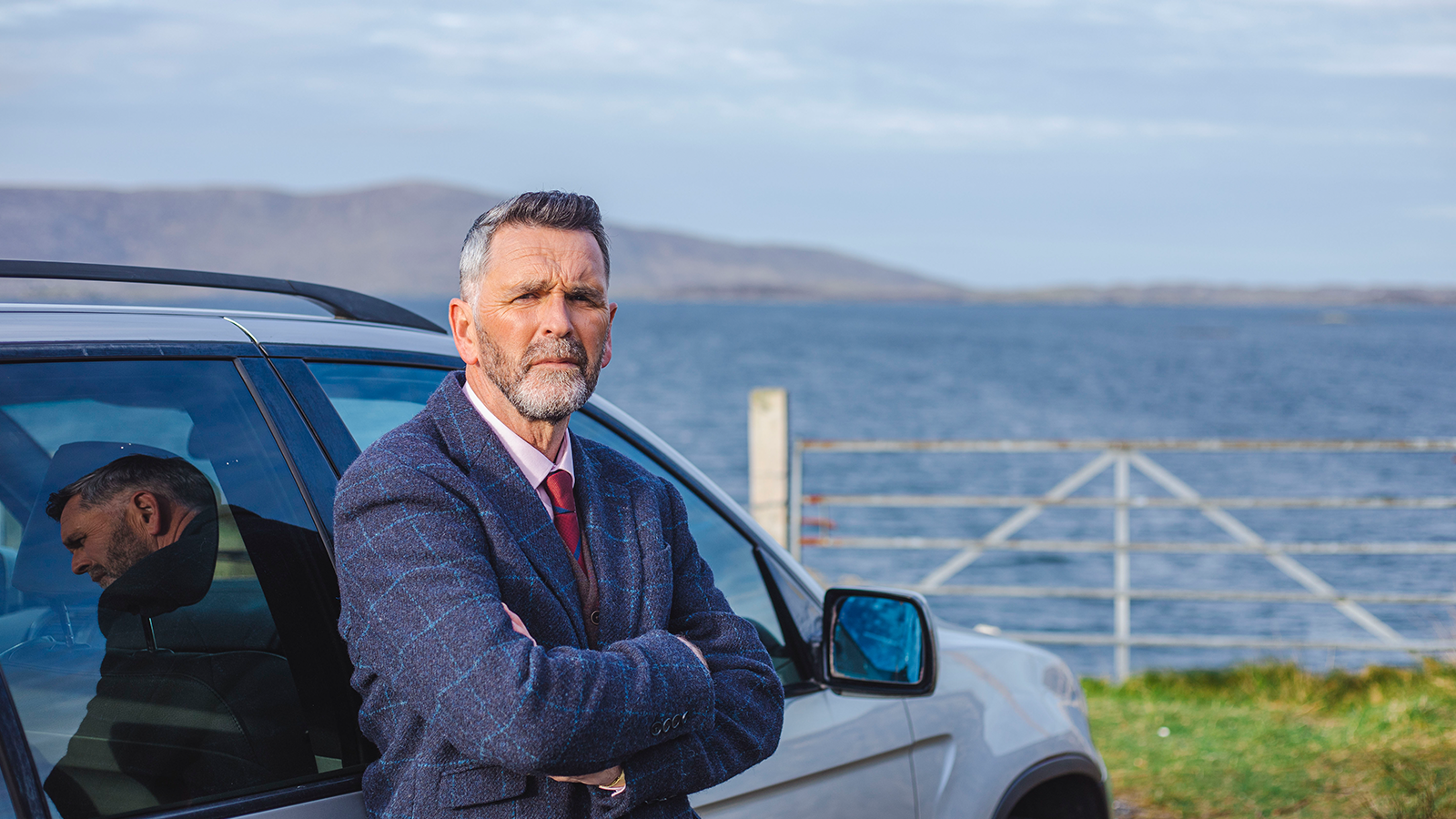 A man in a blue coat leans against a car. In the background is a rusting gate and a large open body of water and mountains in the distance.