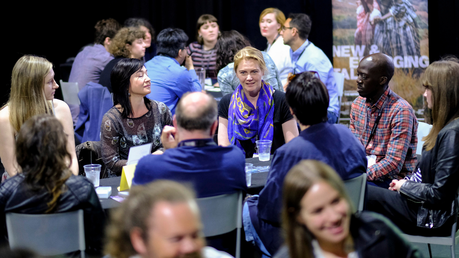 Eight people sit around a table in a group. They are listening to one of the people in the group talking They are surrounded by lots of other people also talking in groups.