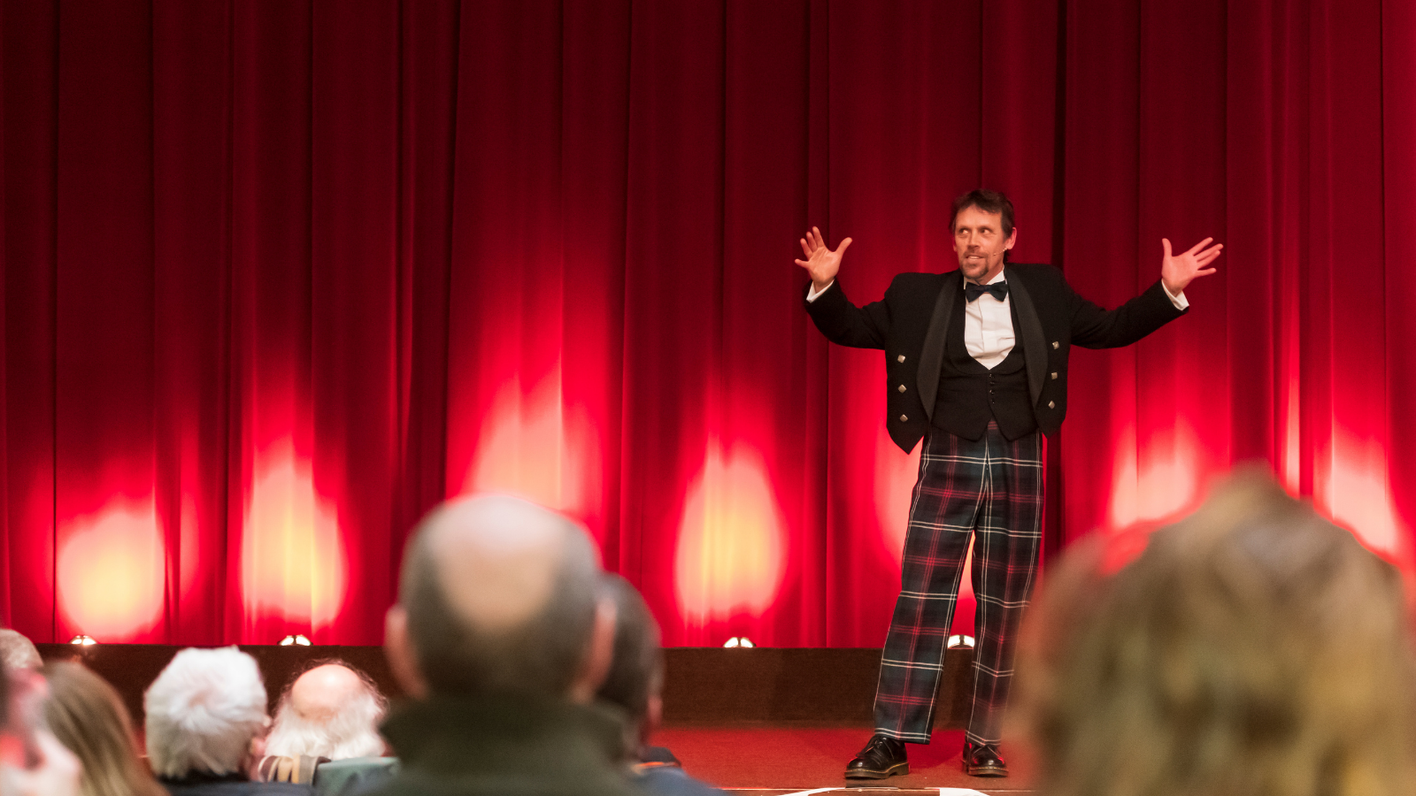 A man in a three piece suit with tartan trousers (Andy Cannon) performs on a stage in front of a pair of closed red curtains. In the foreground are the back of the heads of people watching him.