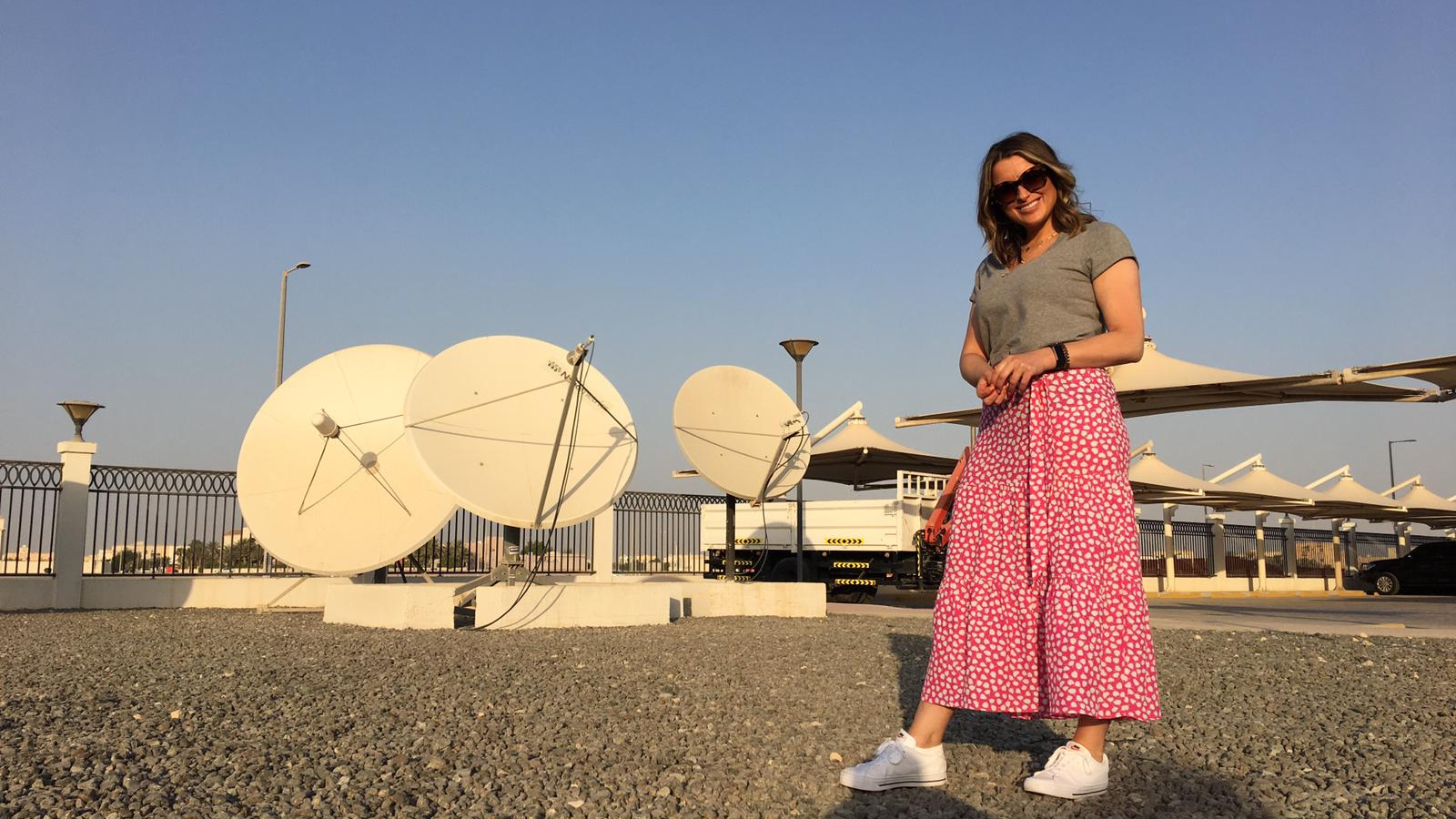 A woman in a red and white skirt and grey top with short blonde hair (Anne McAlpine) stands in a large open area in front of three large white satellites.