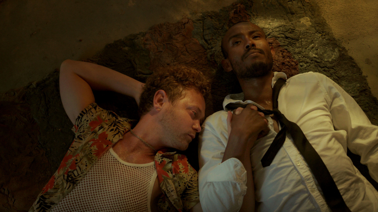 A still from BBQ & Apocalypse. Two men lie next to each other on the floor, holding hands.