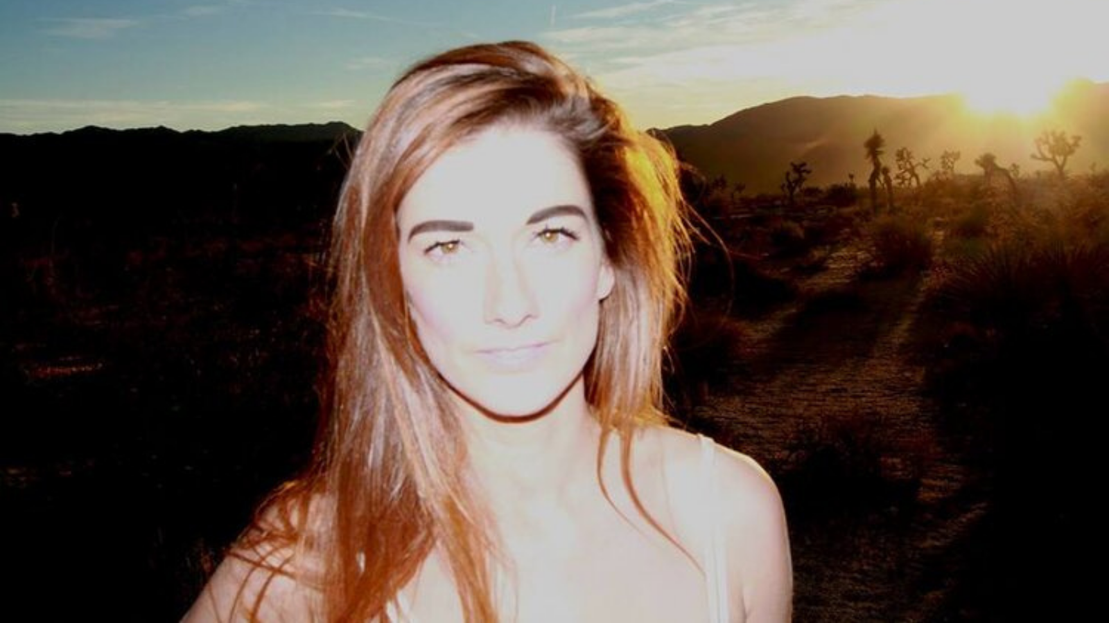 A woman with long reddish brown hair is pictured in bright white light. Behind her the sun peeks out over some hills. This is screen practitioner Mairi Claire Bowser.