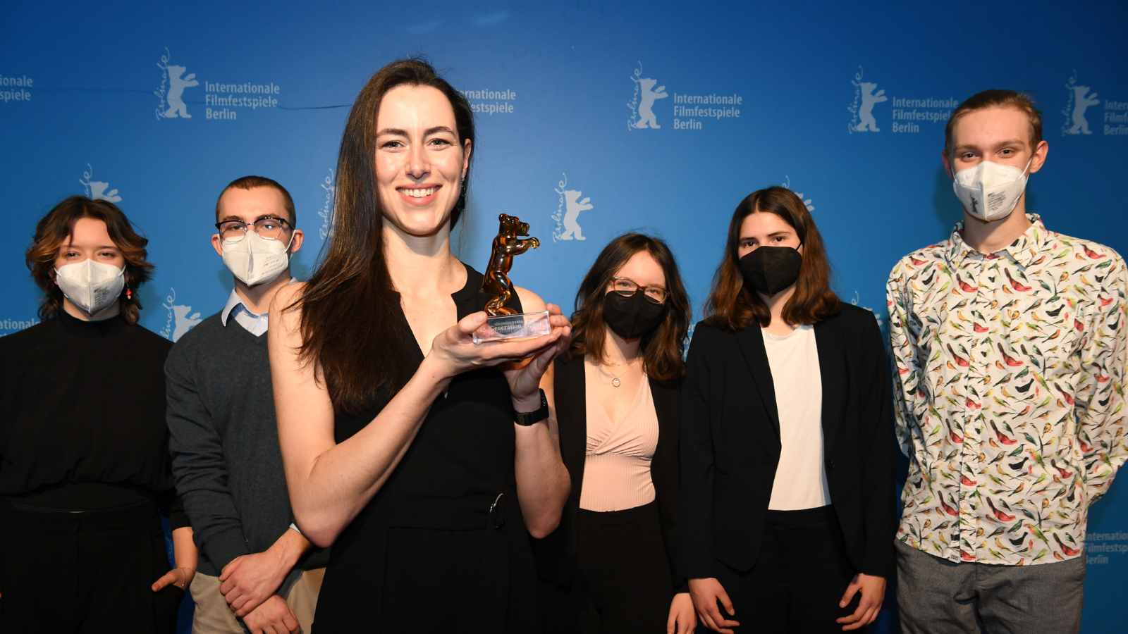 A woman in a black outfit holds an award up, smiling. She is film maker Charlotte Hailstone. Behind her are five people wearing face masks. They are pictured together on a red carpet in front of a bright blue wall, after an awards ceremony.