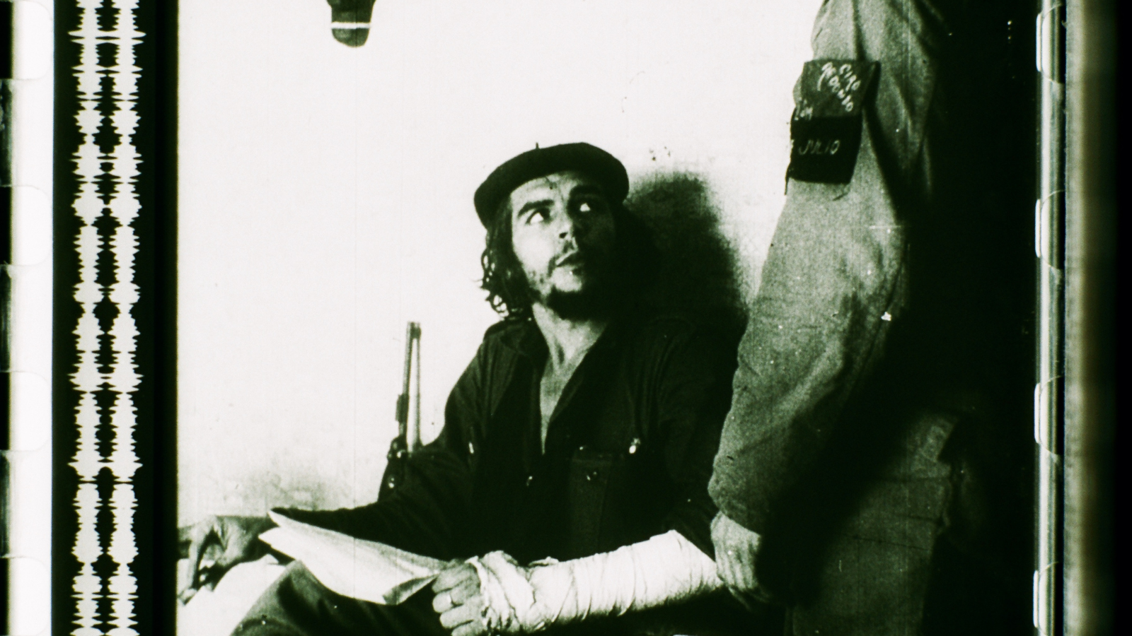 A still from Santiagi Álvarez. It is black and white and shows a man in a beret, seated, looking up at another man in uniform.