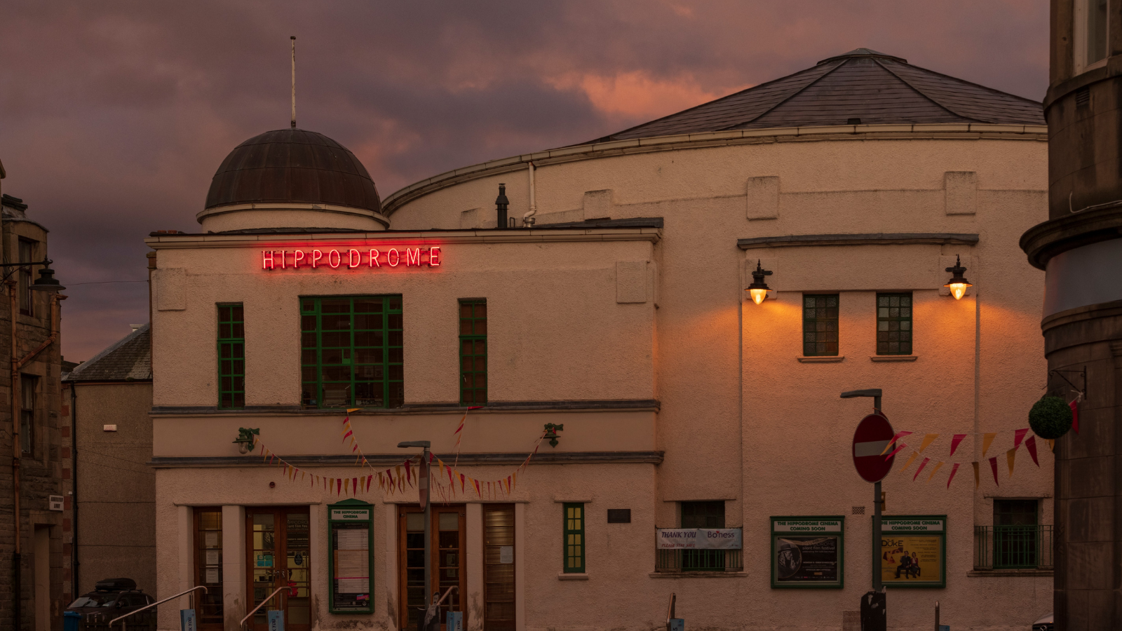 A large 1920s style building at dusk. The word HIPPODROME is lit up in bright red lights.