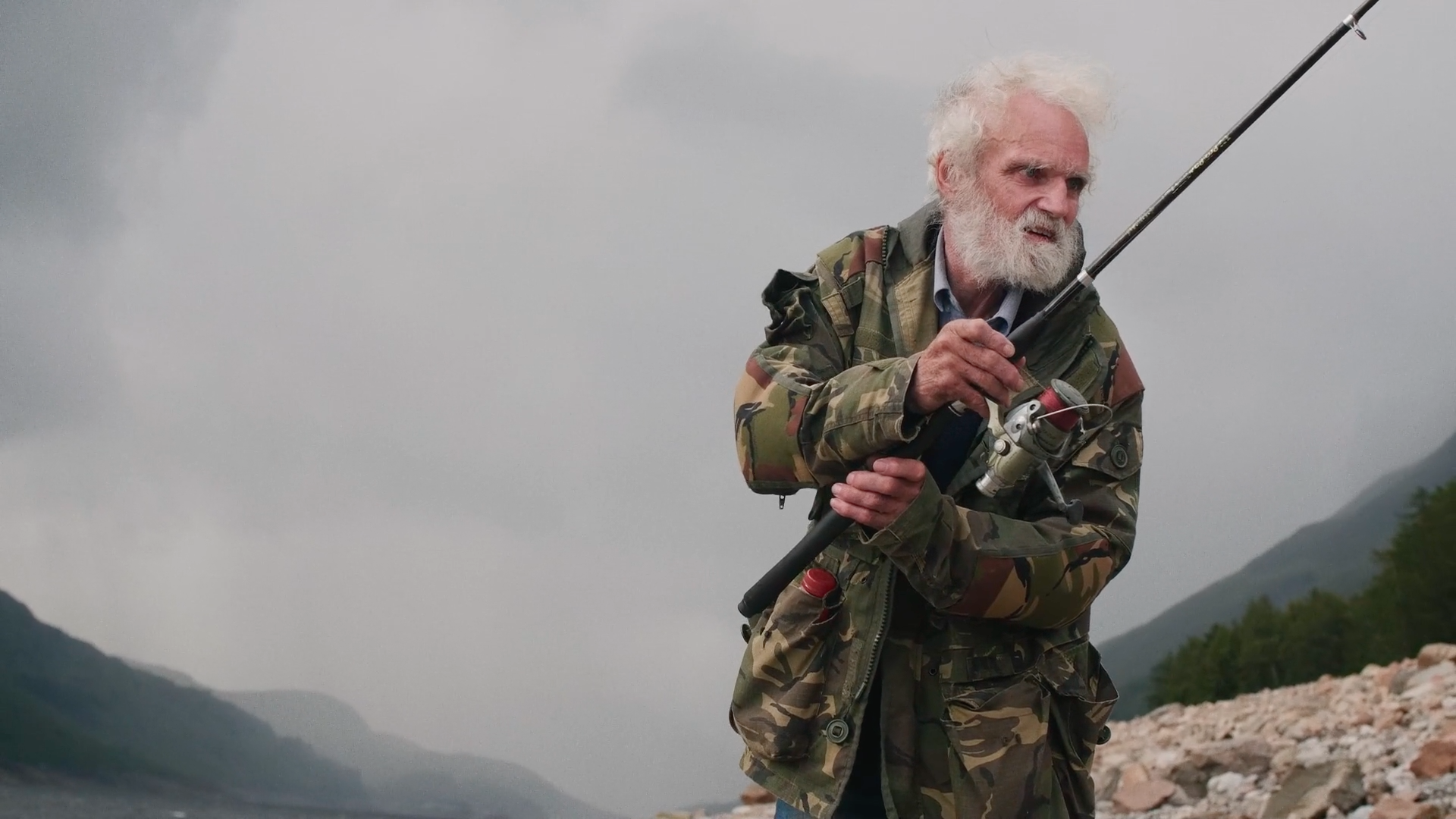 An older man with a white beard and wearing a big camouflage jacket is fishing during a grey overcast day.