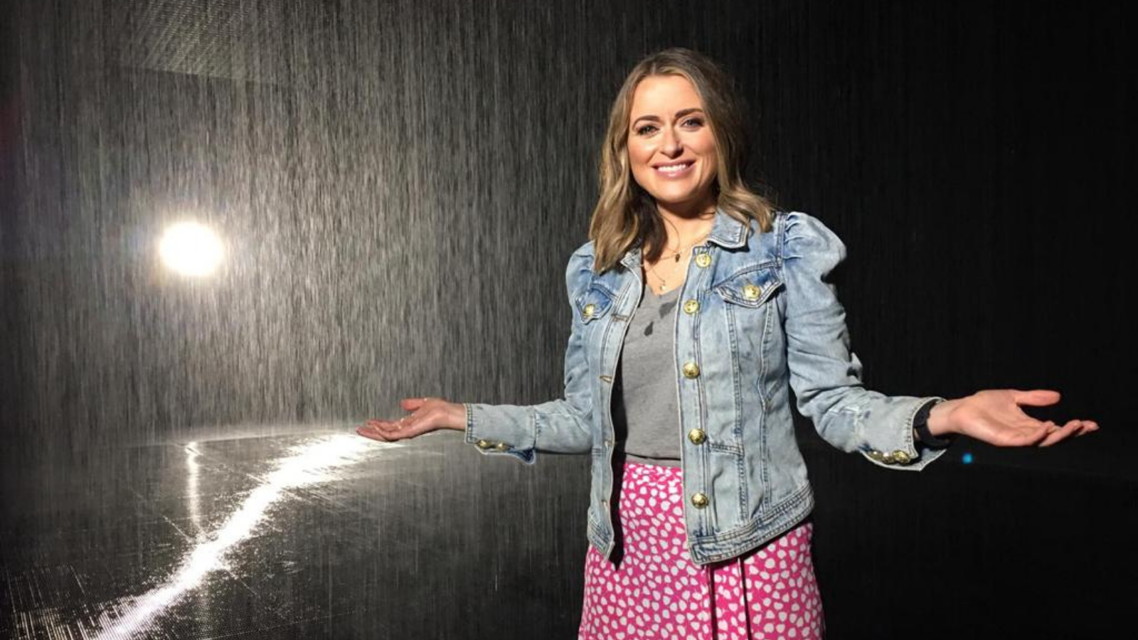 A woman in a red and white skirt, grey top and denim jacket stands with her arms open. She is presenter Anne McAlpine.