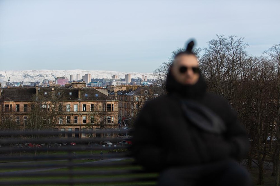 A man in all black - his figure is blurred so as to obscure his features. Behind him are tall Scottish stone tenements. It is winter and the trees have no leaves, and there is snow on the mountains in the background.