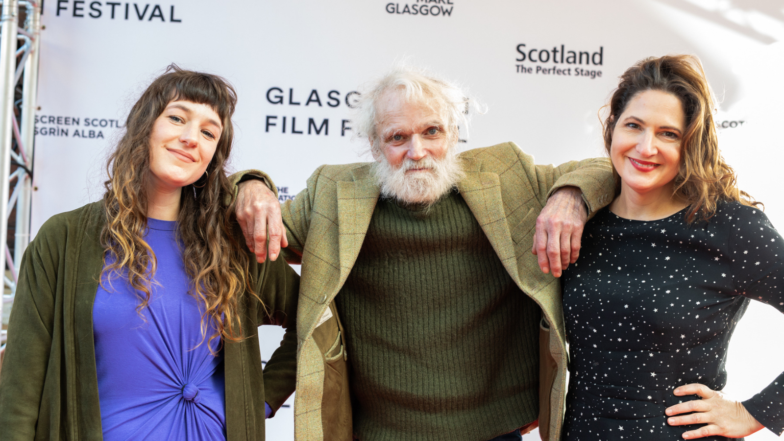 Three people. A man with white hair and a beard who is in the centre, a woman with long brown hair and a fringe on the left, and another on the right who has short curly hair - stand together with their arms around each other.