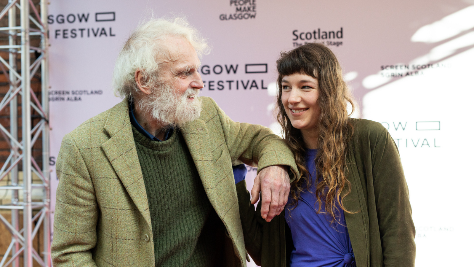 A woman with long brown hair, and an older man with white hair and a white beard (Lizzie MacKenzie and Ken Smith) smile together at a film event