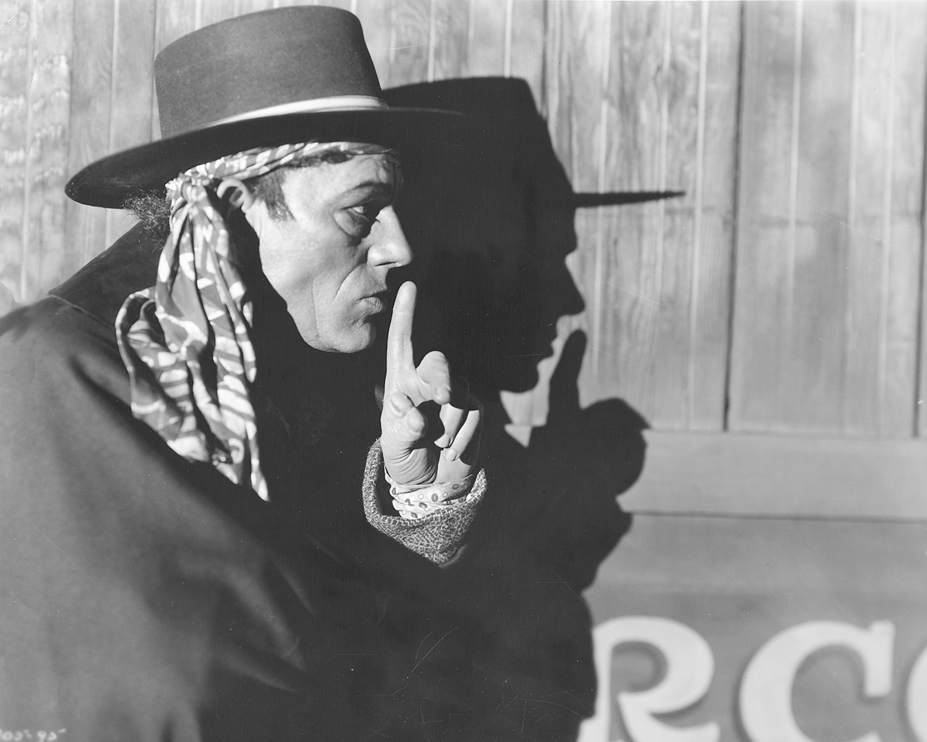 A black and white image. A man in a hat, scarf and cloak holds his finger up in a 'shhhh' motion. It is Lon Chaney in The Unknown (1927) © WBEI