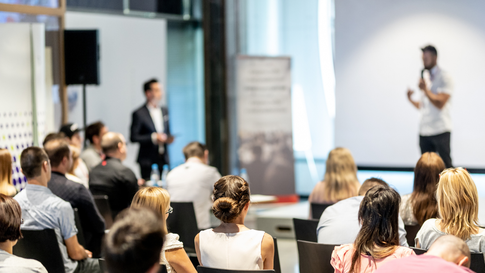 A busy meeting room where a man in a white shirt and black trousers is presenting before a group of professionally dressed people.