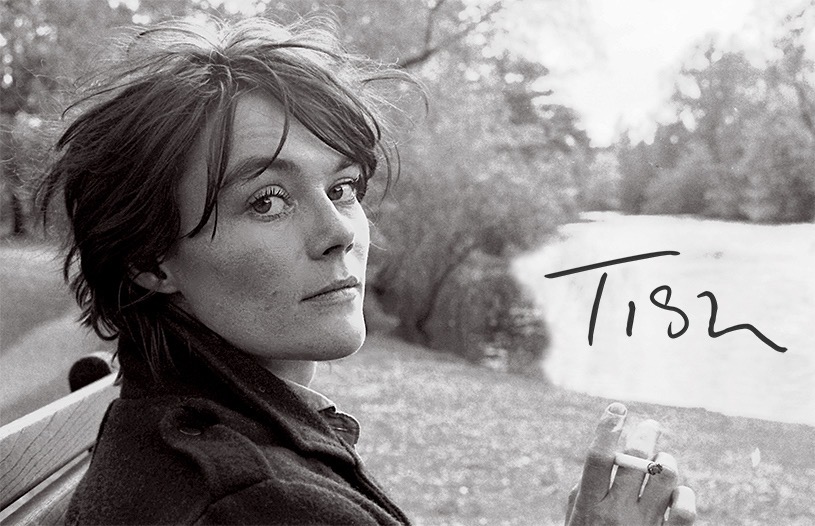 Still from the film TISH, Credit: Ella Murtha. The black and white image shows British documentary photographer Tish Murtha, who is facing away but turning her head to look at the camera. She is in a park by a pond, and holds a cigarette in her right hand. The word Tish is written in black text over the pond.