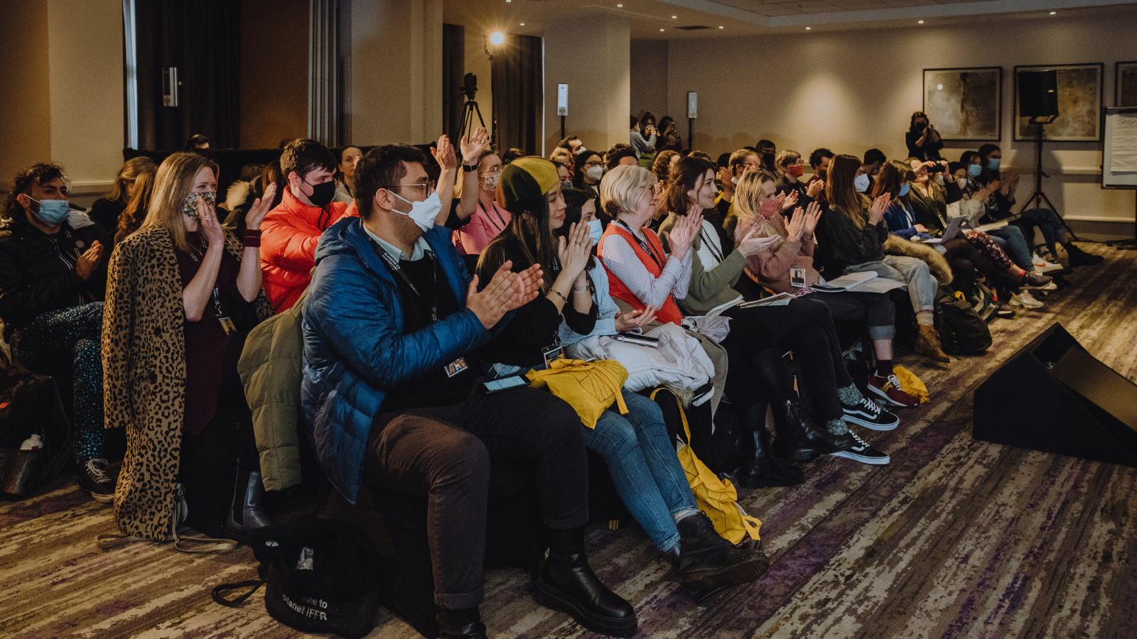 A room full of people sit in rows at a Glasgow Film Festival industry event. There are several long rows, and the people sit clapping as they look to the right of the photo.