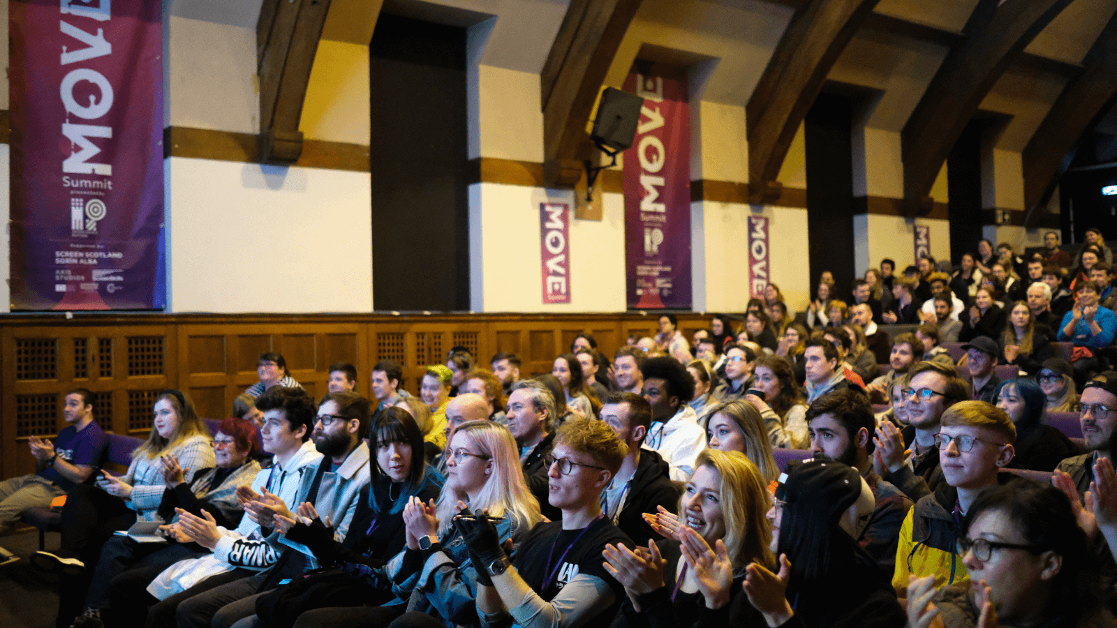 Sat in rows in the University of Edinburgh Pleasance building, an audience at the MOVE summit looks to the left of the image where the stage is off camera, and clap. The audience is a mix of ages and genders, who are clapping.