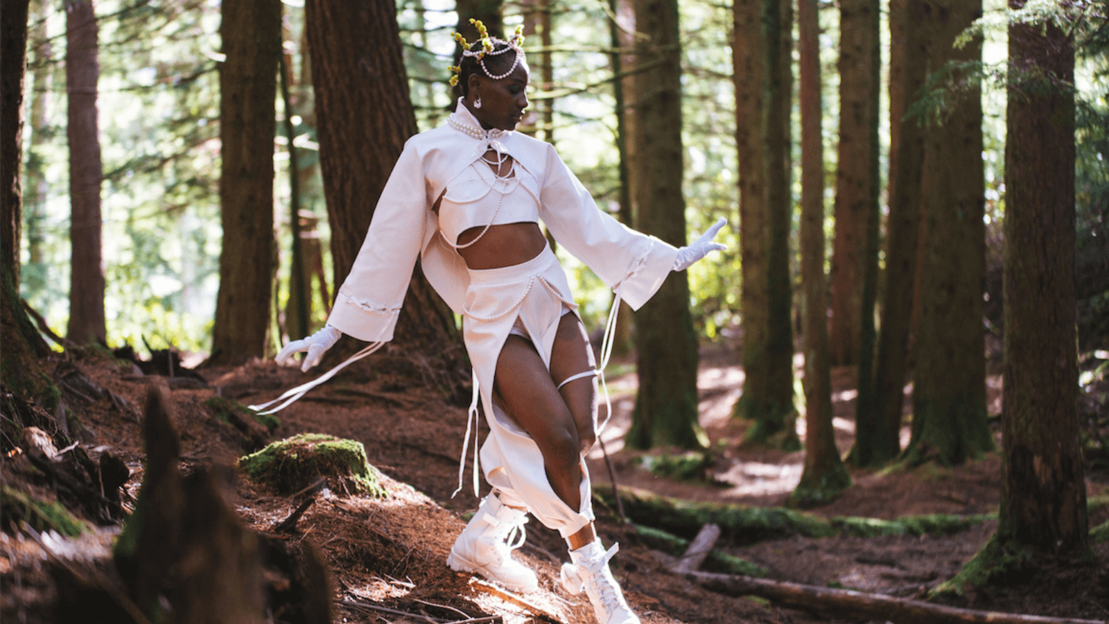 A still from OMOS. Credit: Washington Gwande. Dancer Divine Tasinda stands in a wooded area, wearing a light pink outfit with pearls on it.