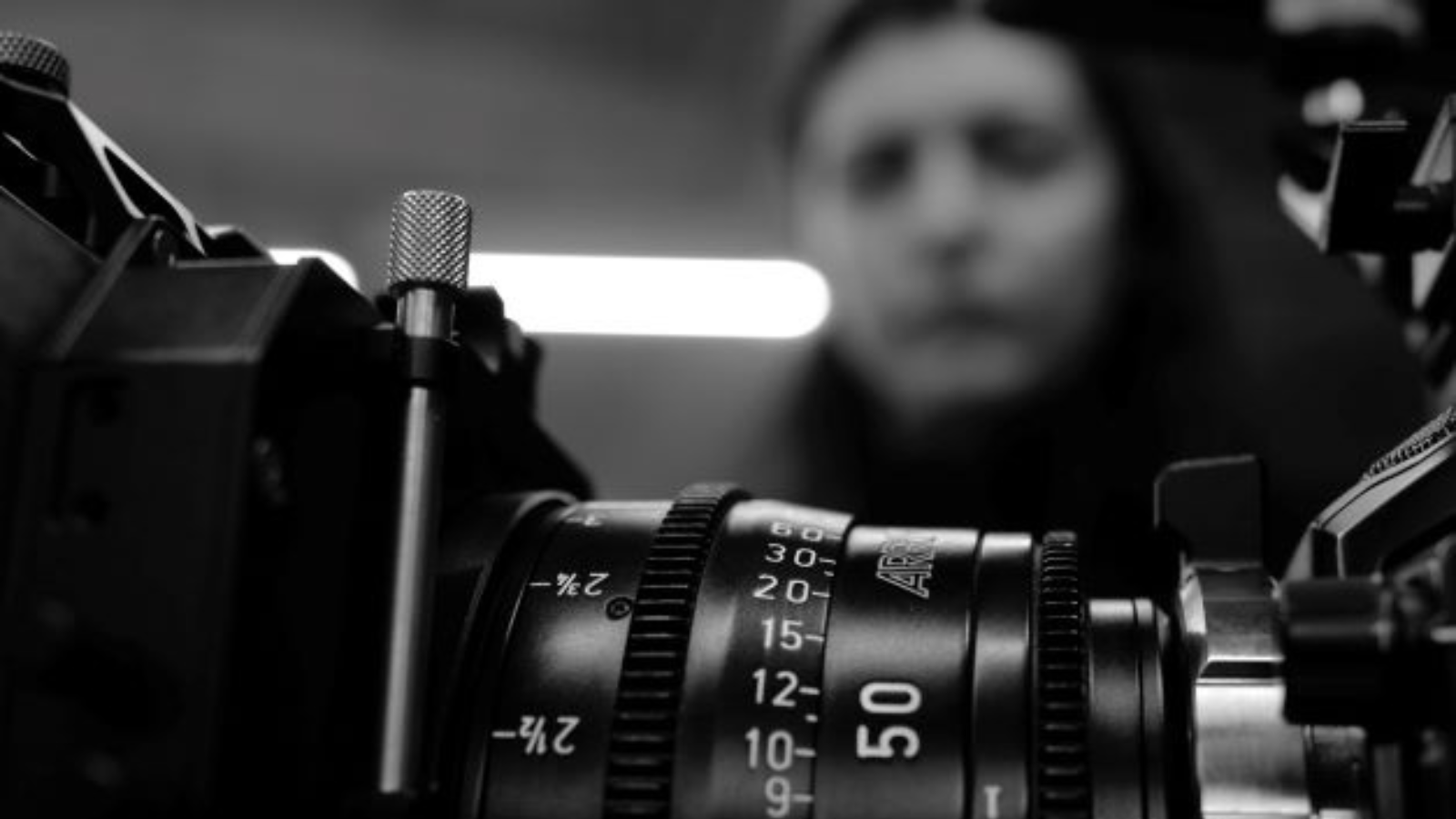 A black and white image which focuses on the lens of a camera in the foreground. In the background and out of focus is the head and shoulders of a woman.