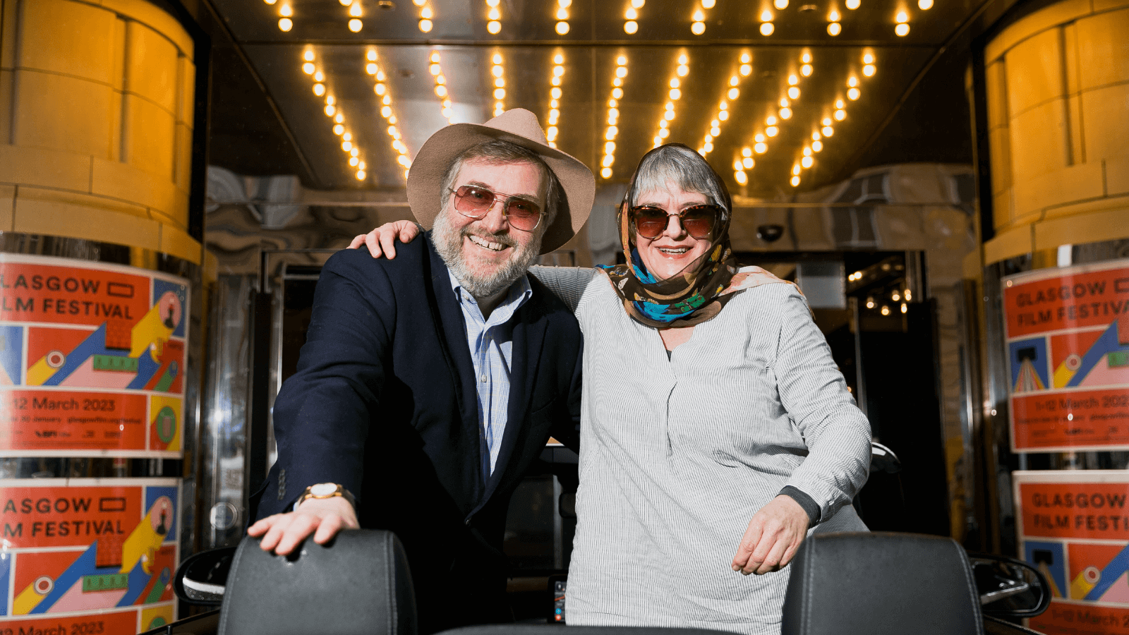 Courtesy of GFF  - This image from the launch of the Glasgow Film Festival 2023 features previous Co-Director of the festival Allan Hunter and current Director Allison Gardner standing together in front of the Glasgow Film Theatre. They look happy and wear sunglasses. Allan wears a suit and a hat, and Allison wears a headscarf wrapped over her head and shoulders.
