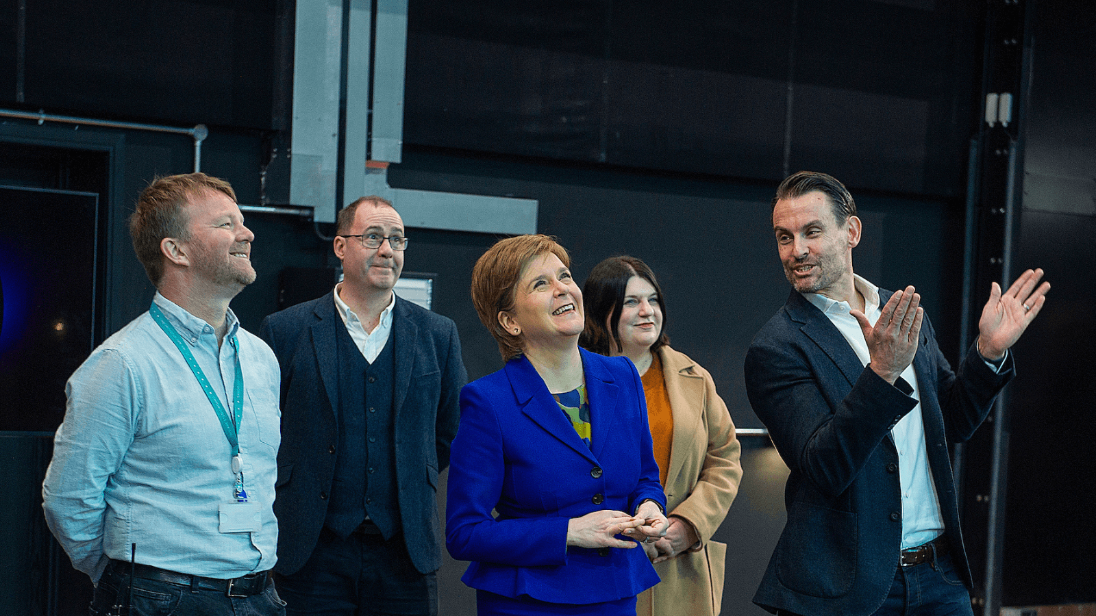 Courtesy of BBC Studioworks - Nicola Sturgeon visits BBC Studioworks, Kelvin Hall. Nicola wears a blue suit jacket and skirt, and is surrounded by two men on her left, including David Smith Director at Screen Scotland, and a man and a woman, Councillor Susan Aitken, Leader of Glasgow City Council on her right. The man on her right wears a suit and is looking at her, while pointing out someone to the top right, which everyone else is looking at.