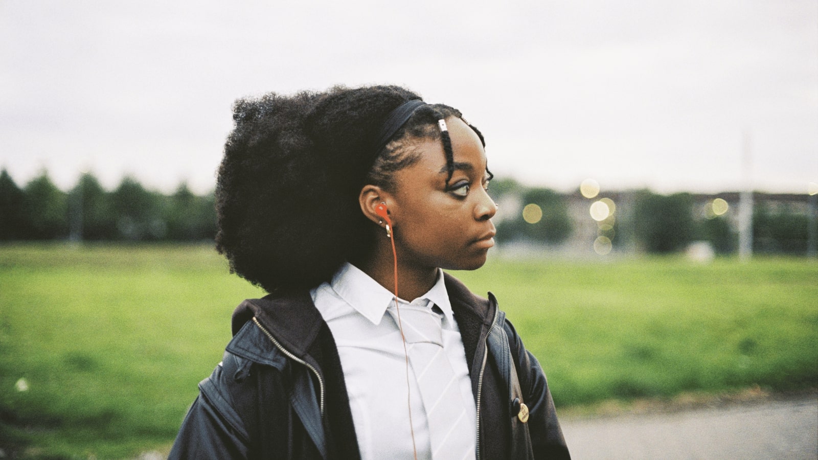Still from short film Blackwool, courtesy of Short Circuit. Actress Miriam Nyarko stands outdoors in a park looking to the right, she wears white school uniform shirt and tie, and a black leather jacket.