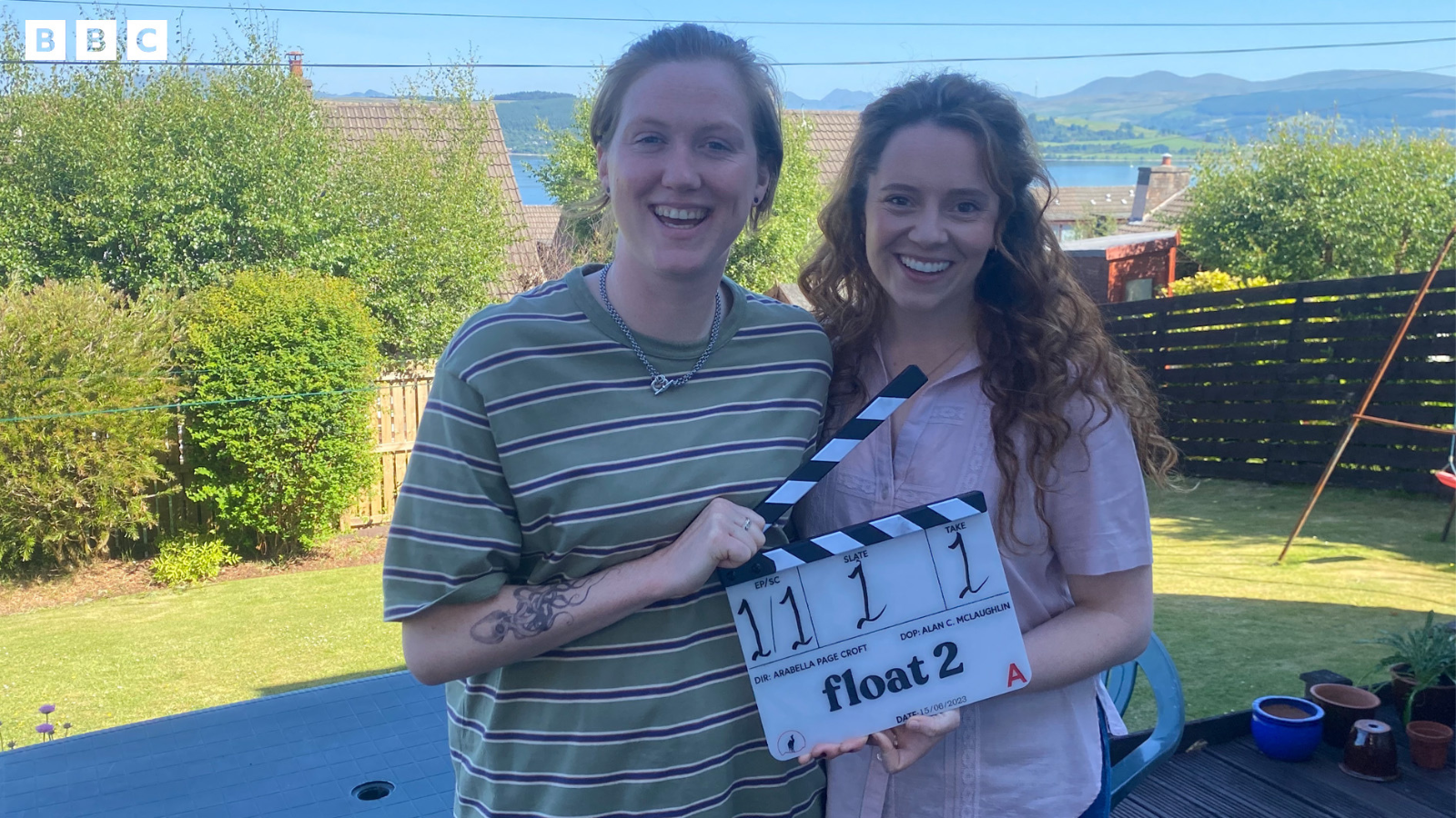 Actresses Hannah Jarrett Scott and Jessica Hardwick stand in a garden with trees and shrubbery side by side holding a clapper board with float 2 written on it in black txt.