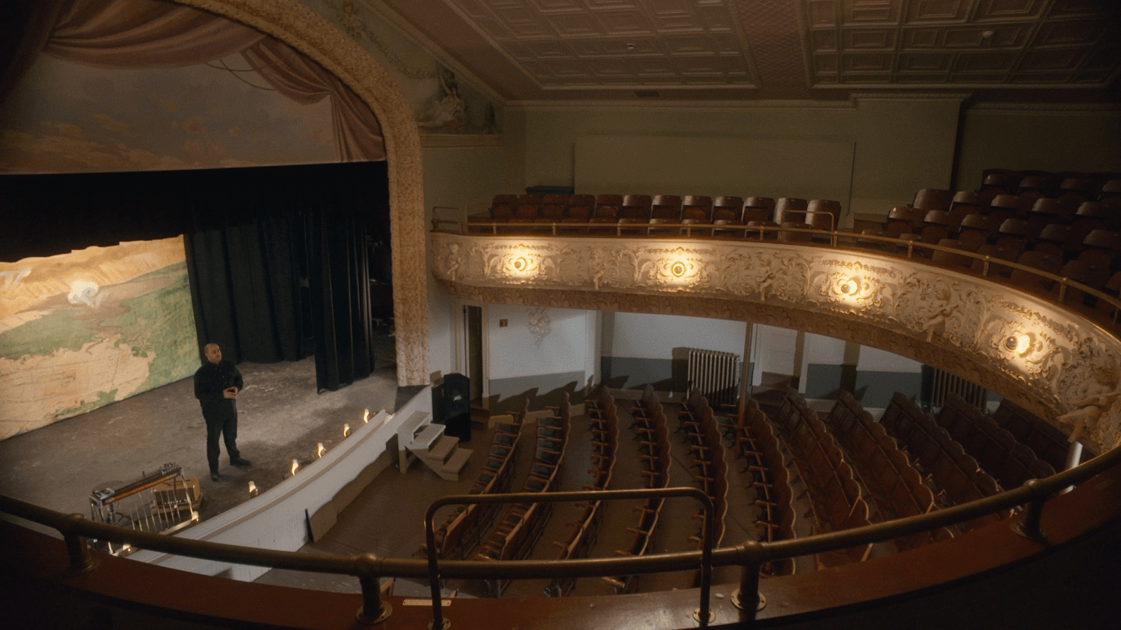 A still from 45th Parallel, courtesy of the Glasgow Short Film Festival. It shows the inside of a theatre, with the stage on the left looking out towards seating on the right of the image. A man wearing black stands on the stage facing the empty seating.