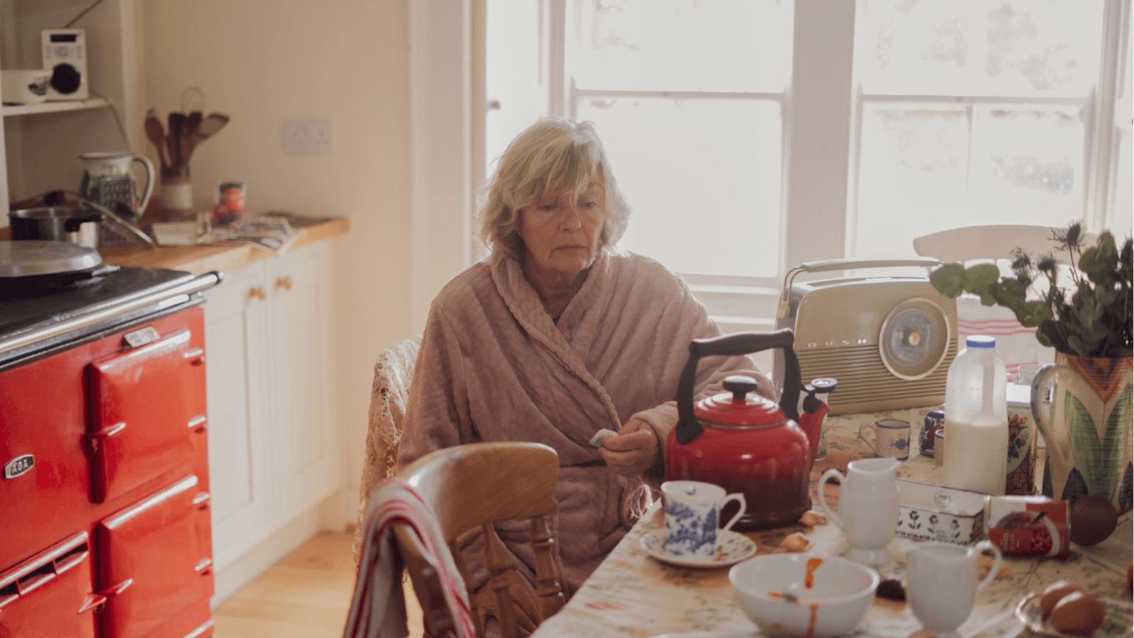 Still from My Name is Beth. Elderly woman in dressing gown sits at messy kitchen table with a red teapot in front of her and solemn expression on her face.