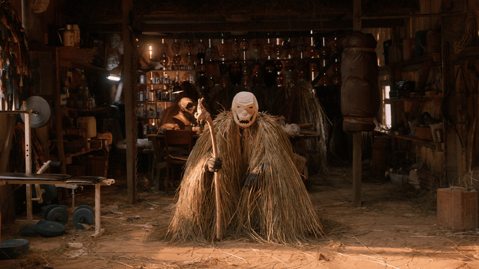 Still from Pamfir. Man in wooden workshop covered in a cape made of dried grass and wearing a white magic which is smiling.