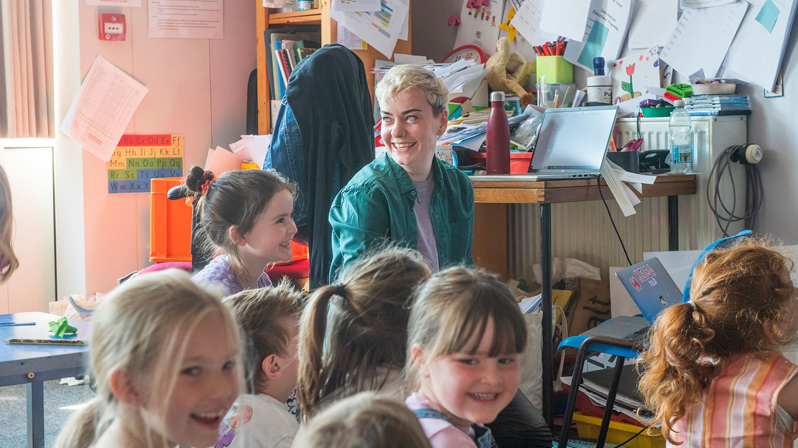 A woman in blue shirt sits amongst a group of young children in a primary school classroom